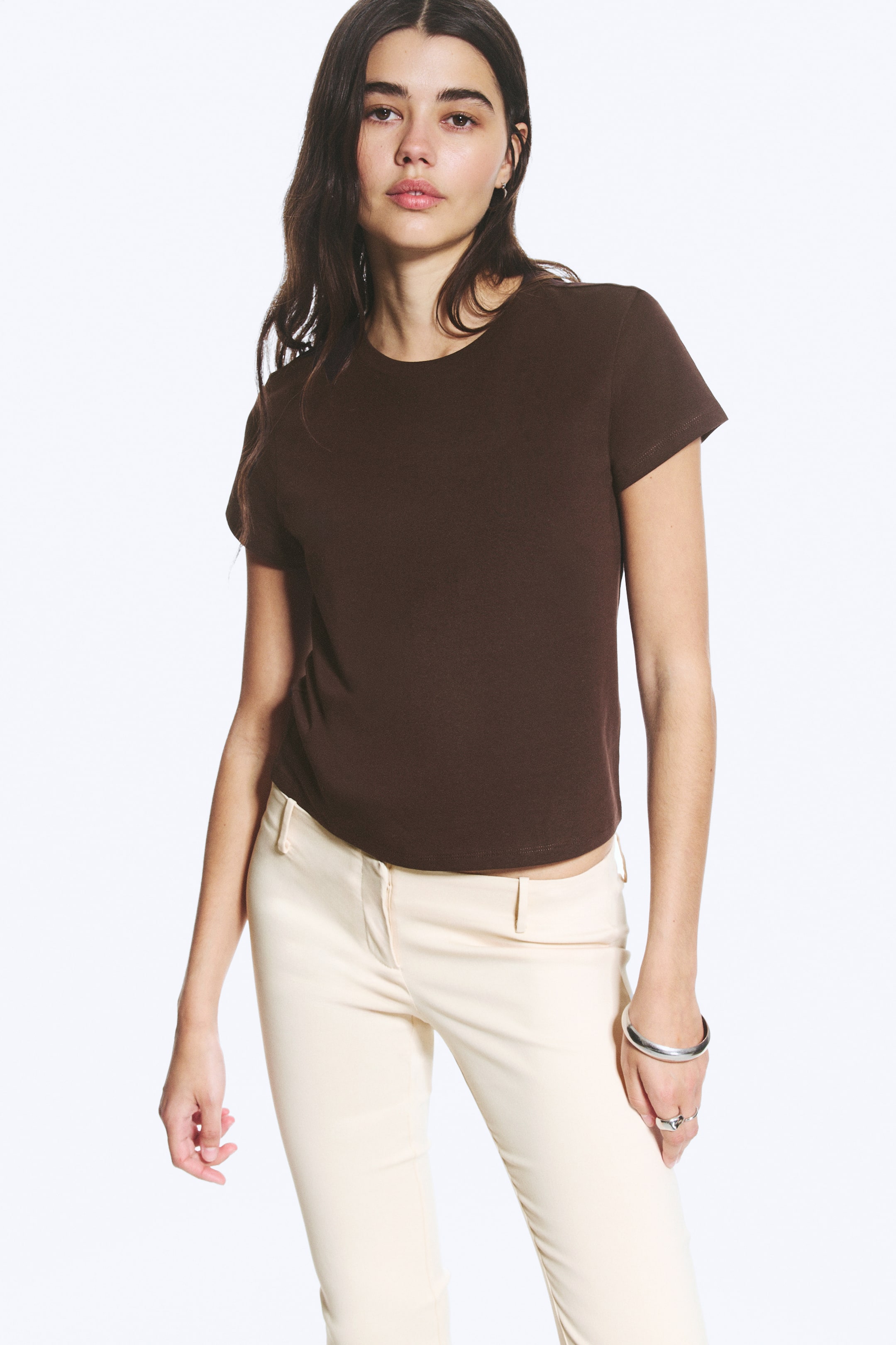 Agrandir l'image: Un mini T-shirt marron foncé à coupe régulière, avec un col rond, des manches courtes à revers et un ourlet droit, est porté par le mannequin. Le T-shirt est associé à un pantalon de couleur claire.