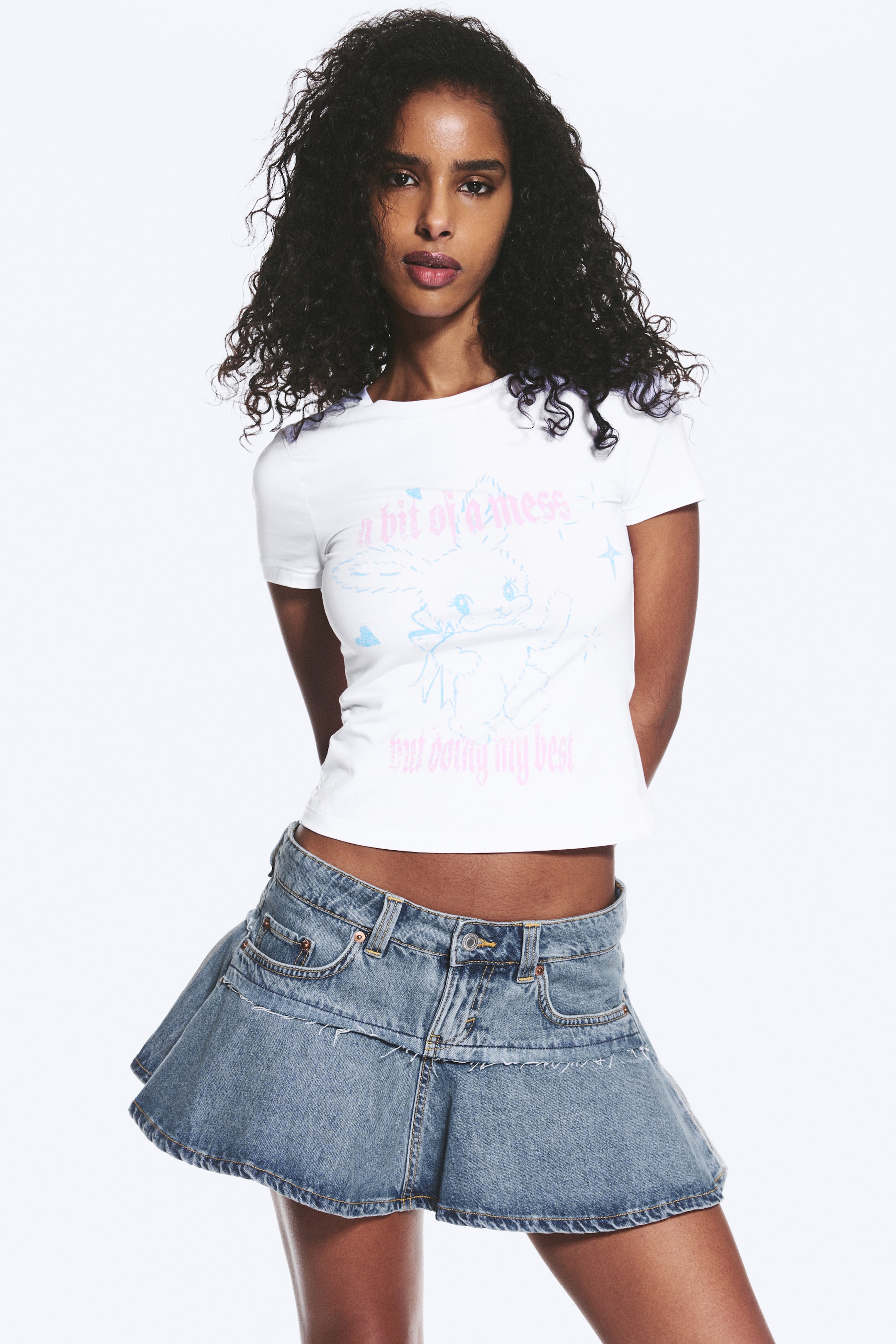 Grotere afbeelding bekijken: Een wit cropped t-shirt met een lichtblauwe cartoonafbeelding van een personage dat een toverstaf vasthoudt, met roze tekst "een beetje een puinhoop" erboven en "maar ik doe mijn best" eronder. Een lichtblauwe denim minirok toont een gelaagd, uitlopend ontwerp met zichtbare naden en voorzakken.