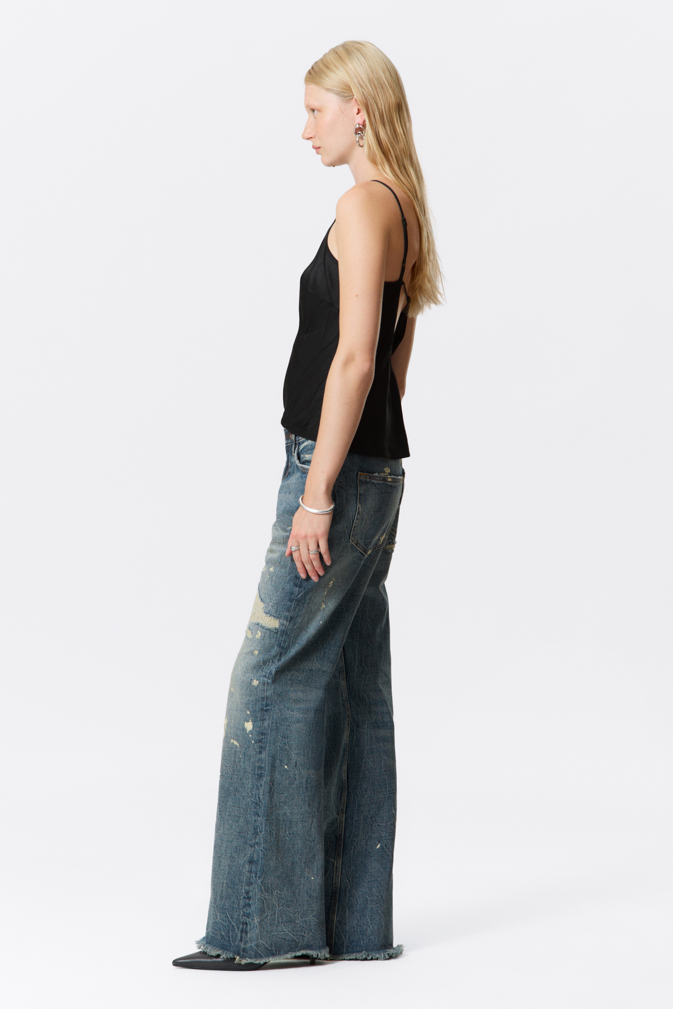 Agrandir l'image: Un jean en denim bleu délavé avec un bas évasé et des ourlets bruts est porté par le mannequin. Le jean présente des abrasions importantes et des marques plus claires à l'avant, et est assorti d'un débardeur noir et de chaussures noires à bouts pointus.