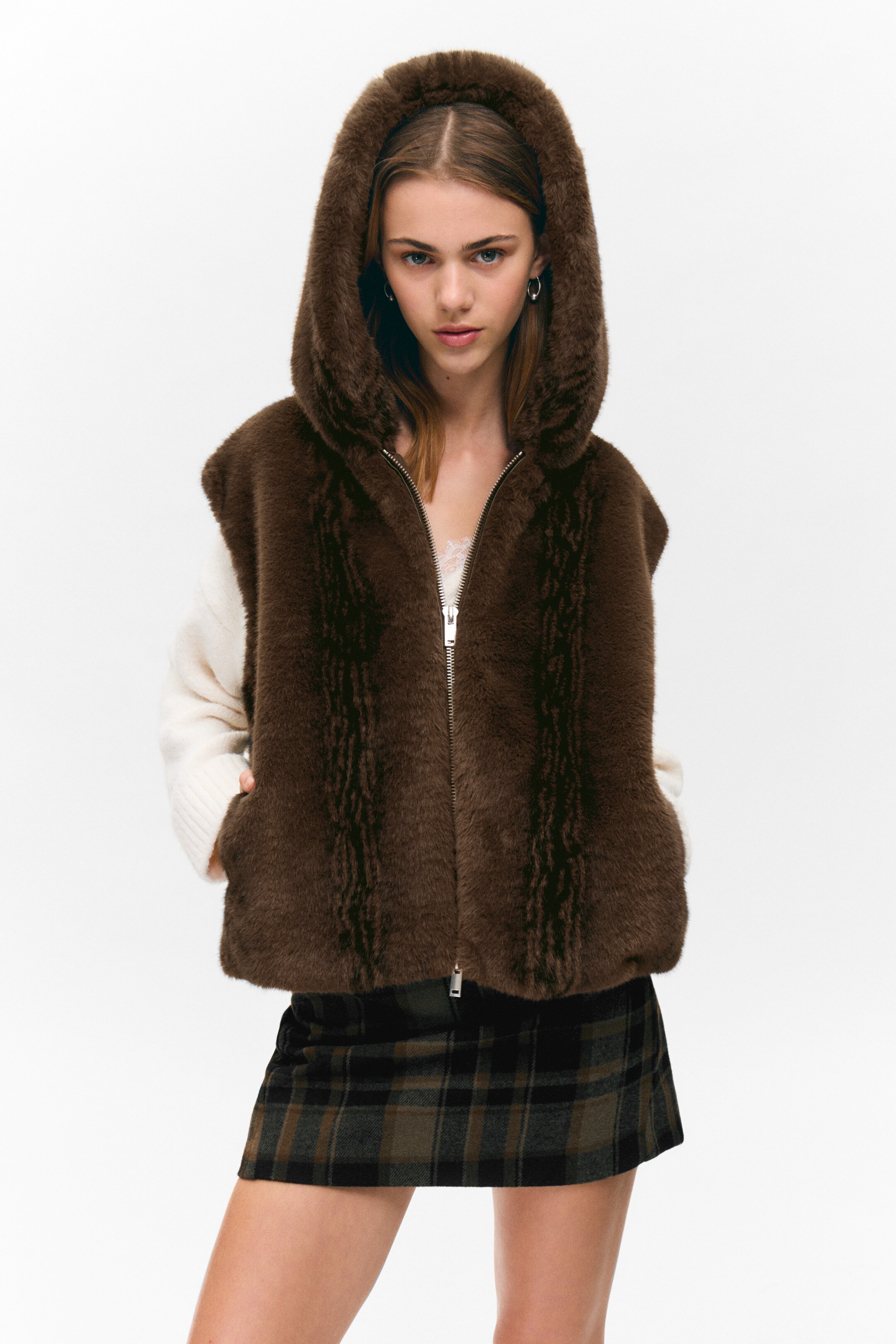 Grotere afbeelding bekijken: Een bruine faux fur vest met capuchon wordt gedragen, naar voren gericht. Het heeft een ritssluiting aan de voorkant, subtiele verticale textuurstrepen op de panelen en zichtbare steekzakken. De vest is gelaagd over een witte trui met lange mouwen en een donkergroene en bruine geruite minirok.