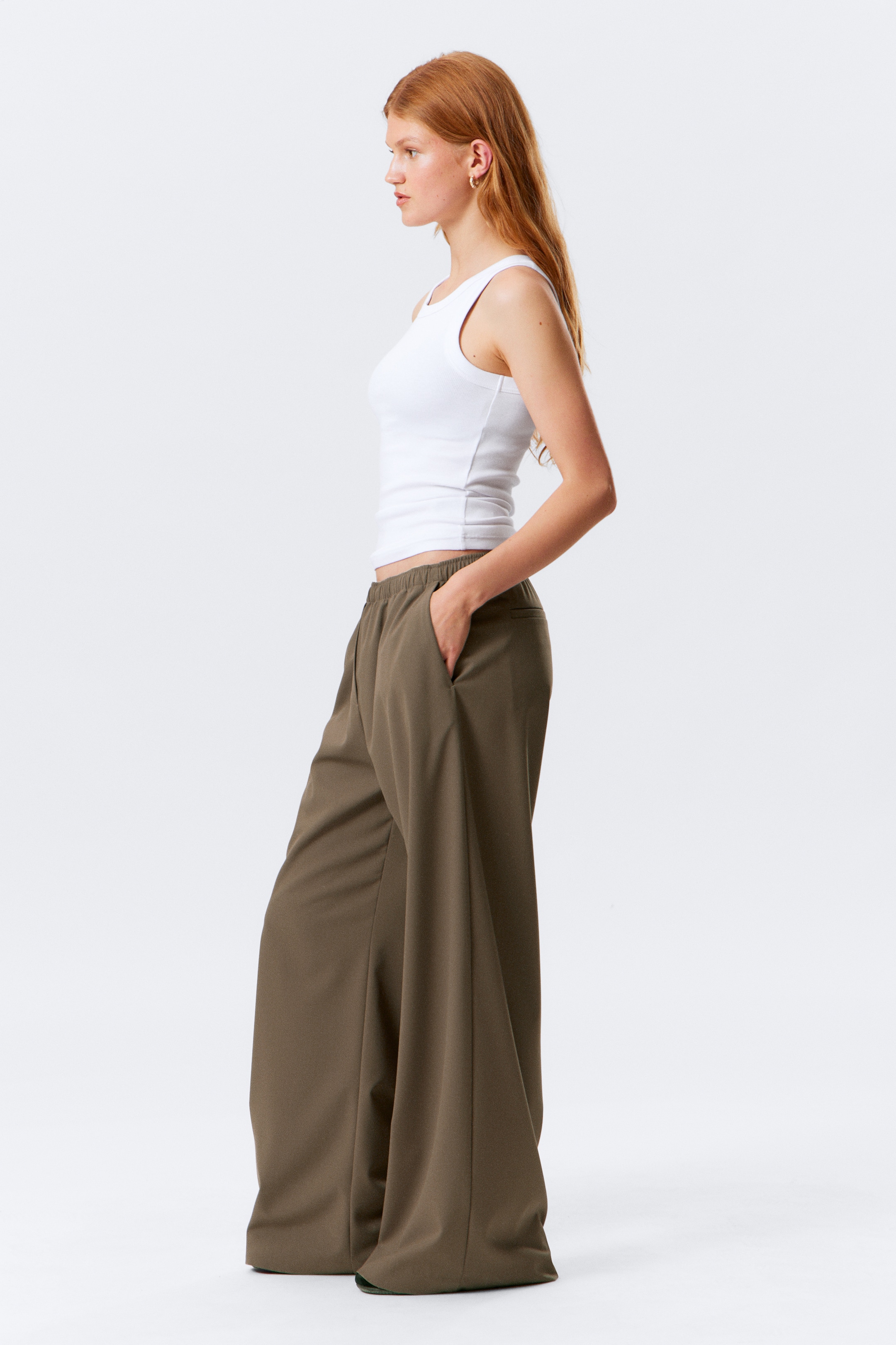 Grösseres Bild ansehen: Ein weißes geripptes Tanktop wird mit olivgrünen, weit geschnittenen Hosen getragen, die in einer Seitenansicht präsentiert werden. Die Hosen haben einen elastischen Bund und sammeln sich an den Füßen, während das Tanktop eng anliegt und an der Taille endet.