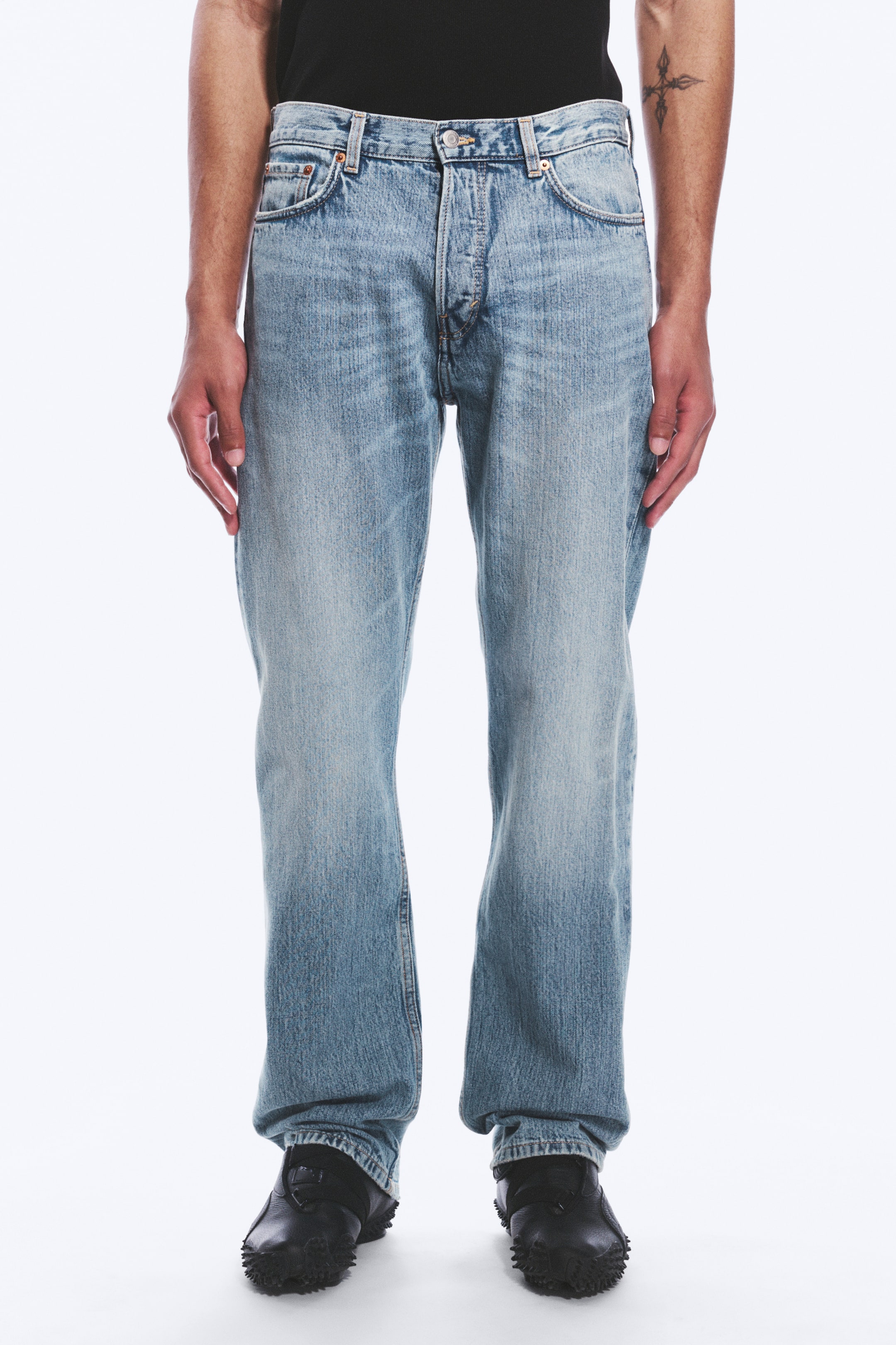 Nagyobb kép megtekintése: A modell közepes derekú, világoskék, merev farmeranyagú farmert visel, amely laza szabású és egyenes szárú. A farmer gombos sliccel és elülső zsebekkel rendelkezik, a nadrágszárak pedig fekete cipőkre támaszkodnak.