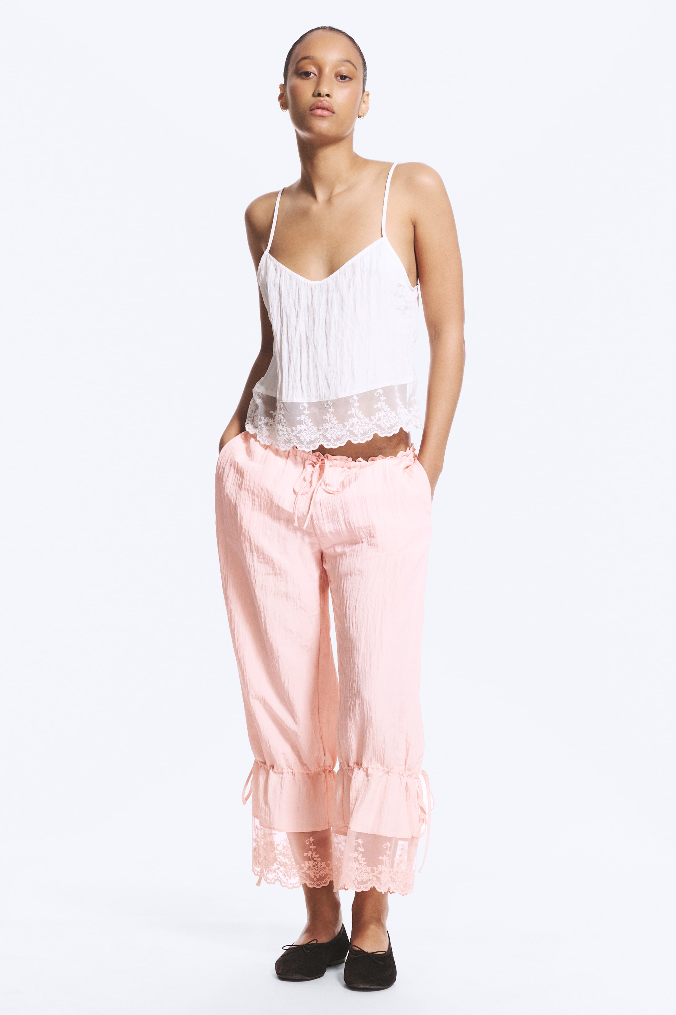 Agrandir l'image: La mannequin porte des bloomers roses taille basse avec une texture froissée, se tenant avec les mains dans les poches latérales. Les bloomers présentent une taille élastique avec cordon de serrage et des jambes trois-quarts avec des ourlets froncés ornés de dentelle.