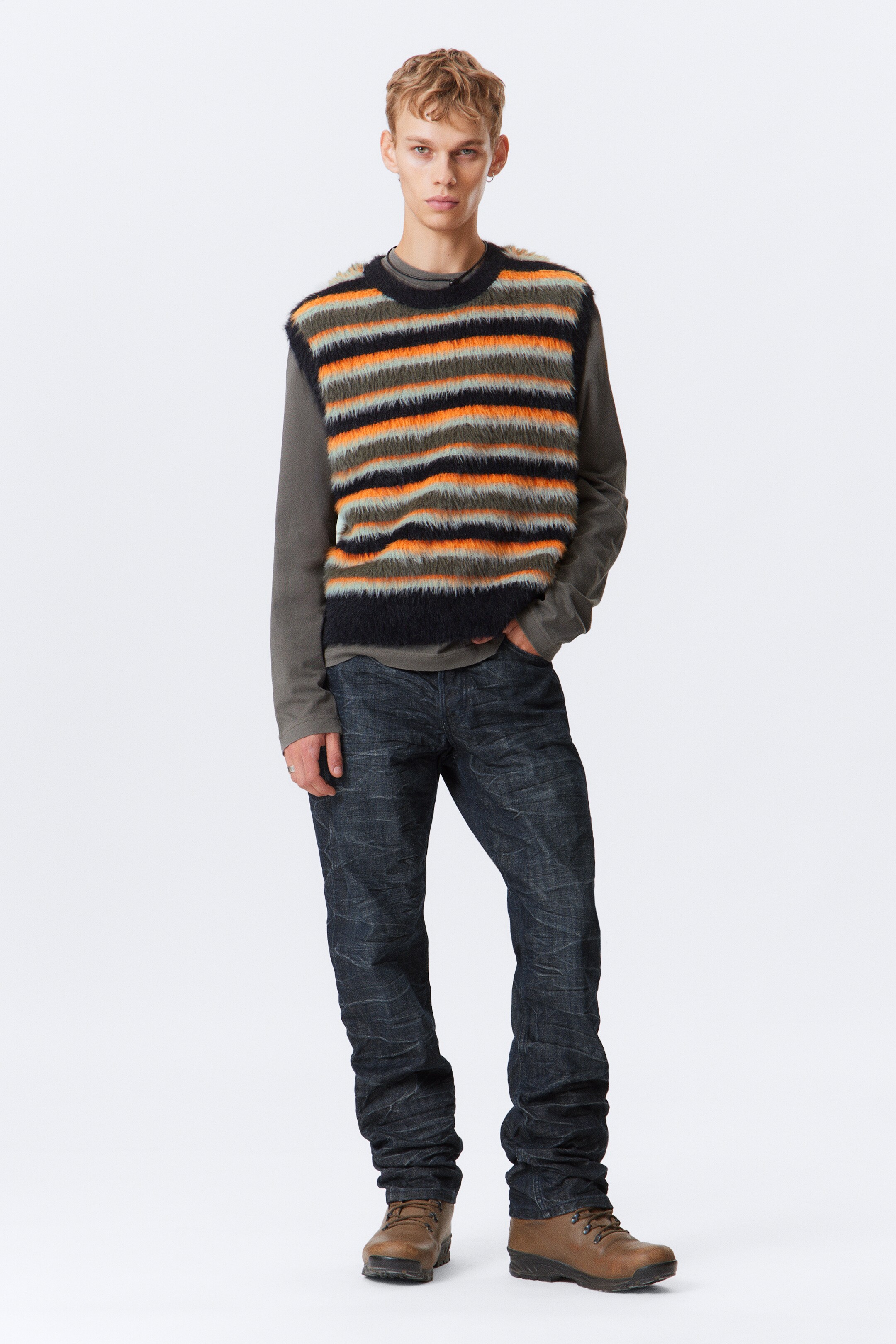 Agrandir l'image: Un gilet pull moelleux, avec des rayures horizontales en noir, orange et vert olive, est porté par-dessus une chemise à manches longues vert olive foncé. Un jean délavé foncé, avec une coupe ample et une texture froissée, s'accumule sur des bottines marron à lacets.