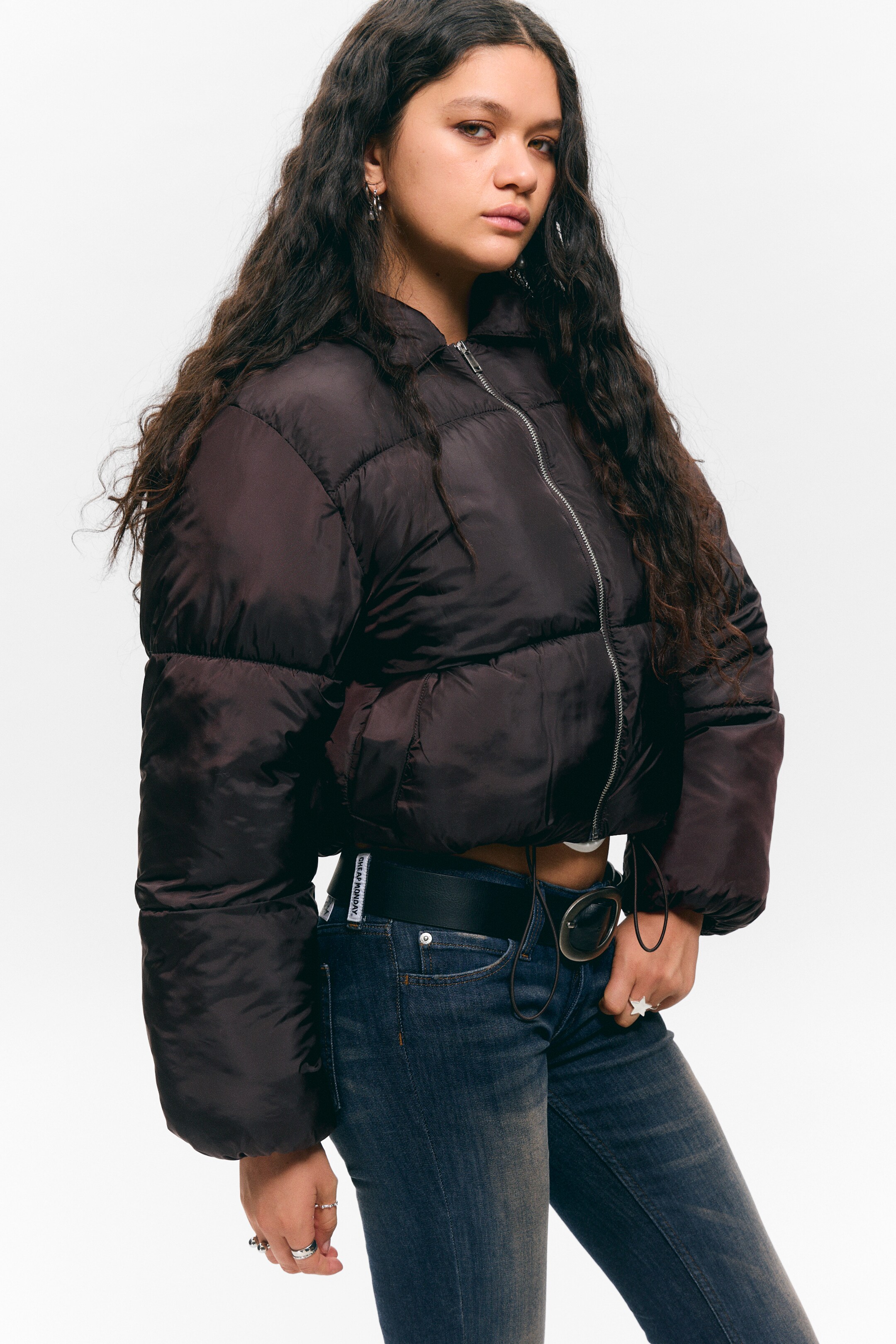 View larger image: A dark brown, cropped puffer jacket is being presented, angled slightly to the right, showcasing its voluminous design. It is featuring a full-length silver zipper up the front to a stand-up collar, and a visible drawstring is cinching the hem on the right side.