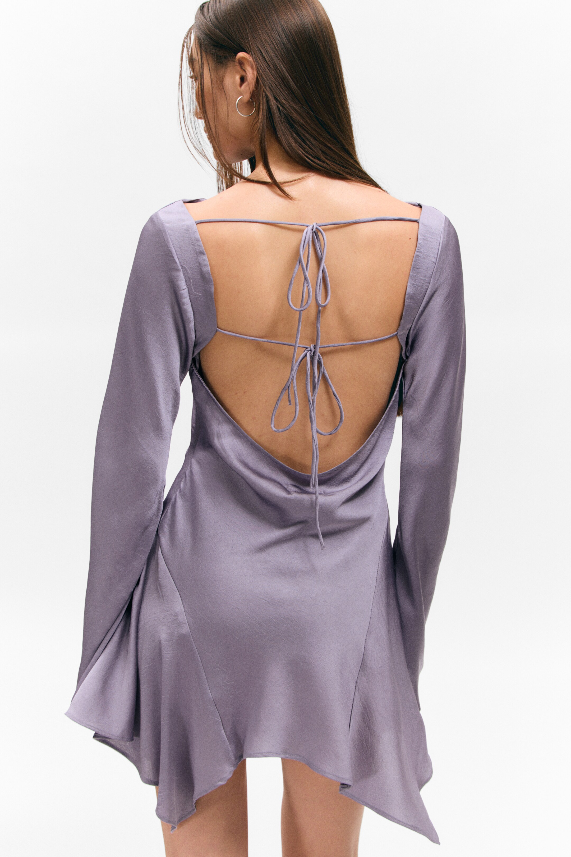 View larger image: A lilac satin-like dress is displaying its back, featuring a deep open-back design with thin self-fabric ties crisscrossing and knotting at the upper back. It is showcasing long sleeves and an asymmetrical draped hem.