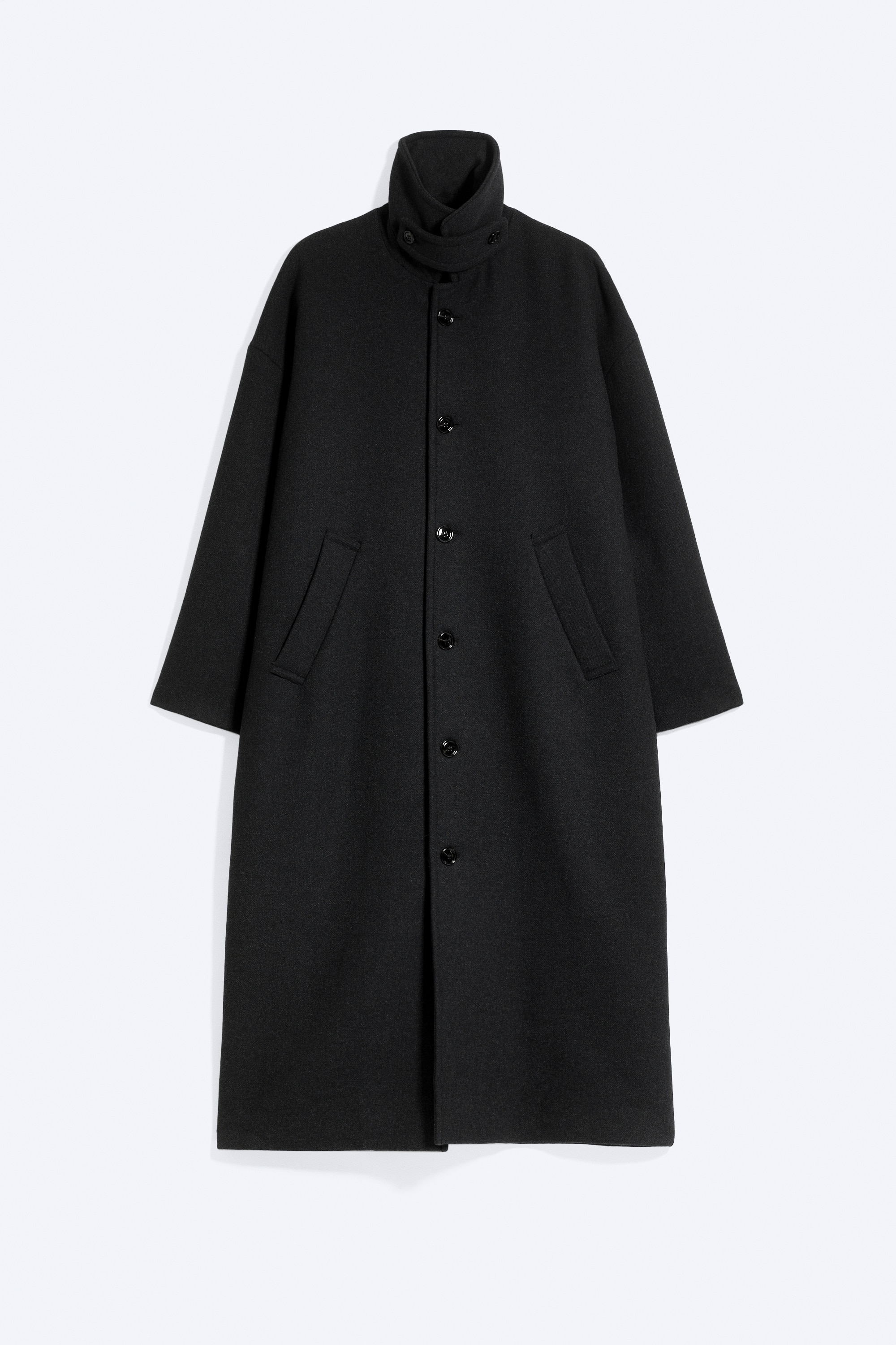 Un long manteau noir oversize est portÃ© par le mannequin, dotÃ© dâun col montant boutonnÃ© et dâune silhouette allongÃ©e. Le mannequin marche de profil, une main glissÃ©e dans une poche passepoilÃ©e en biais, et associe le manteau Ã  un jean bleu dÃ©lavÃ© et Ã  des bottes en suÃ¨de beige.