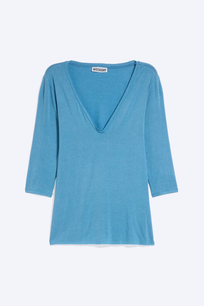 A light blue, three-quarter sleeve top is shown front-facing, featuring a deep plunge V-neckline and a straight hem. The lightweight jersey fabric shows a subtle heathered texture, with a white brand label visible on the inside collar. - 1