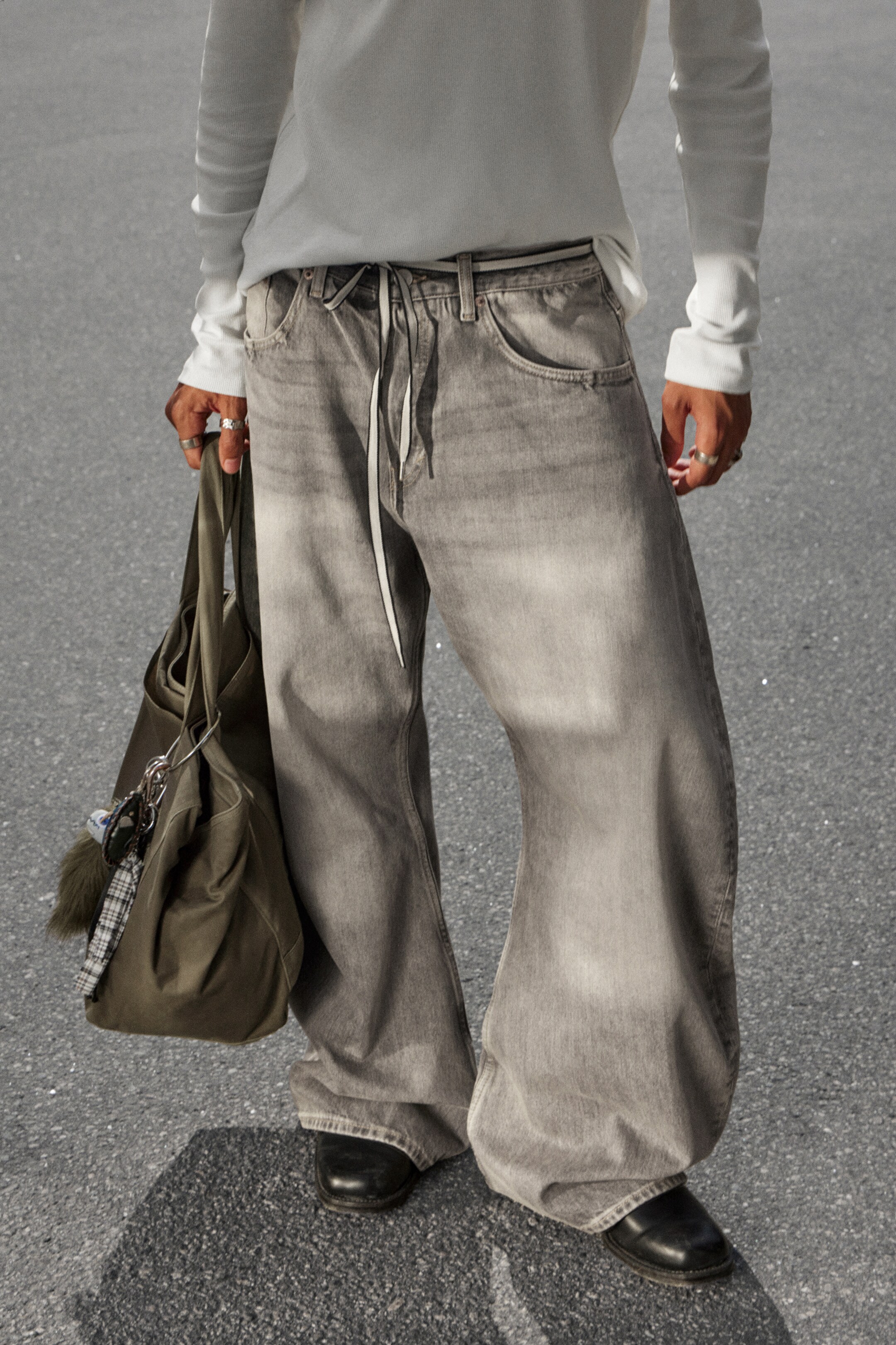 Agrandir l'image: Un pantalon en denim gris est présenté de face, mettant en valeur une coupe très ample et large avec des jambes qui s'accumulent sur les chaussures du porteur. Le pantalon présente un délavage gris délavé, des poches avant inclinées, des passants de ceinture visibles et un cordon de serrage de couleur claire à la taille avant.