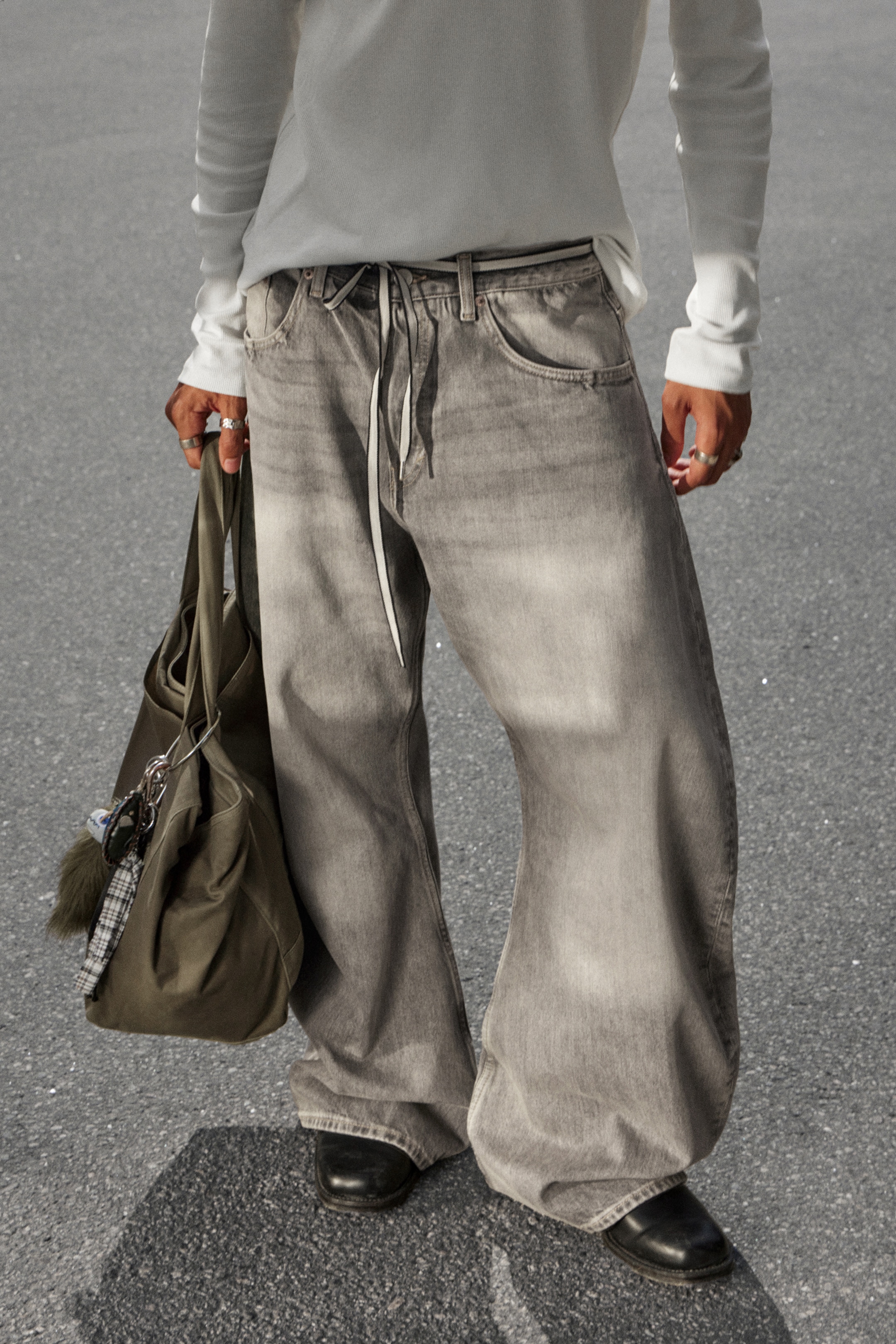 View larger image: Gray denim pants are being presented from the front, showcasing a very loose, baggy fit with wide legs that are pooling over the wearer's shoes. The pants are featuring a faded gray wash, slanted front pockets, visible belt loops, and a light-colored drawstring at the front waistband.