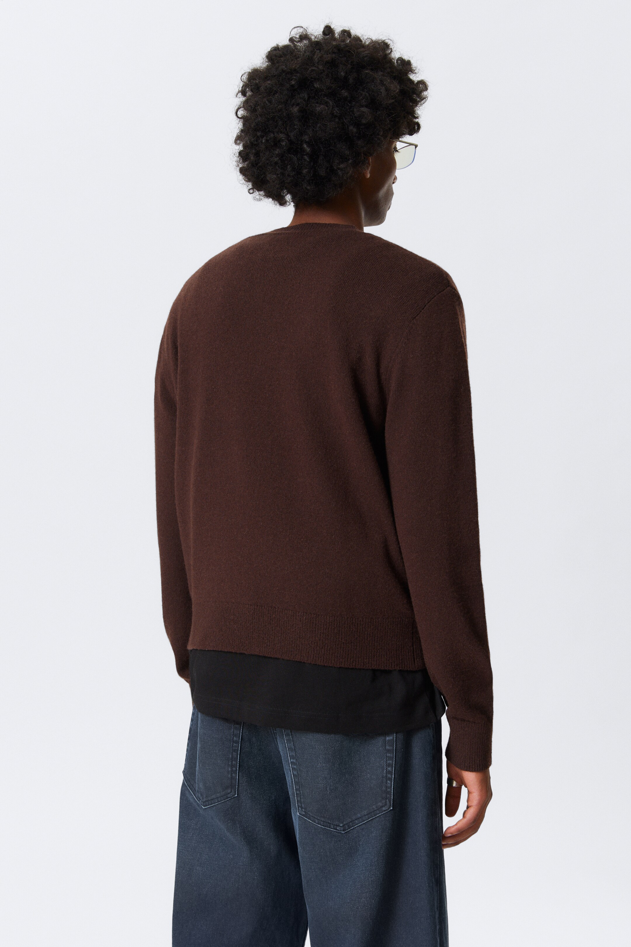Grotere afbeelding bekijken: Een donkerbruine, gladgebreide trui wordt van de achterkant gepresenteerd. Het is ontworpen met lange mouwen en een geribde zoom, en er wordt een zwart t-shirt onder gedragen, waarvan de zoom onder de trui uitsteekt.