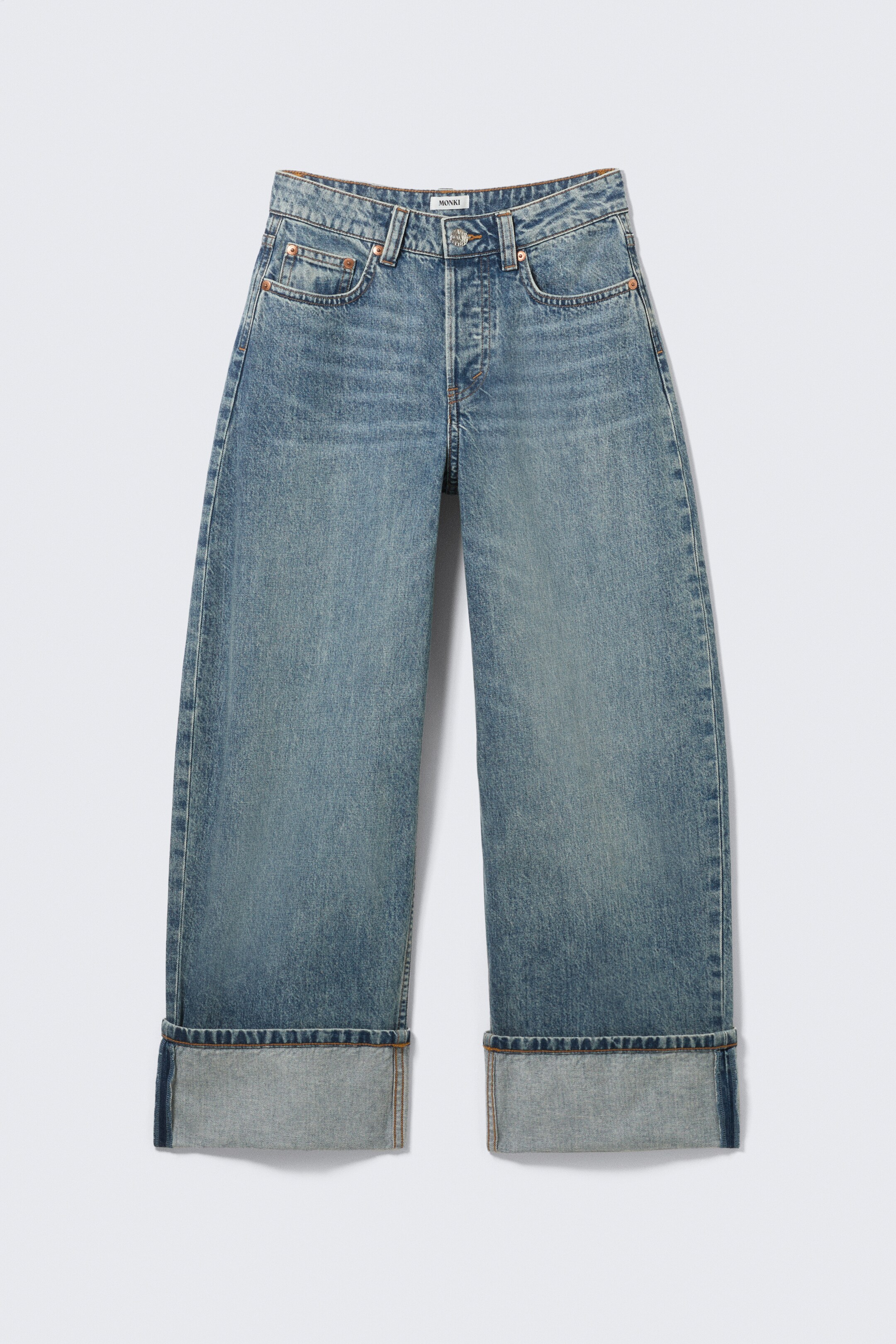 Visa större bild: Ett par medelljus tvättade blå jeans med låg midja presenteras FRAM, med en lös passform och breda, bylsiga ben. De visar upp en klassisk femficksdesign, en knappgylf och stora manschetter vid anklarna.