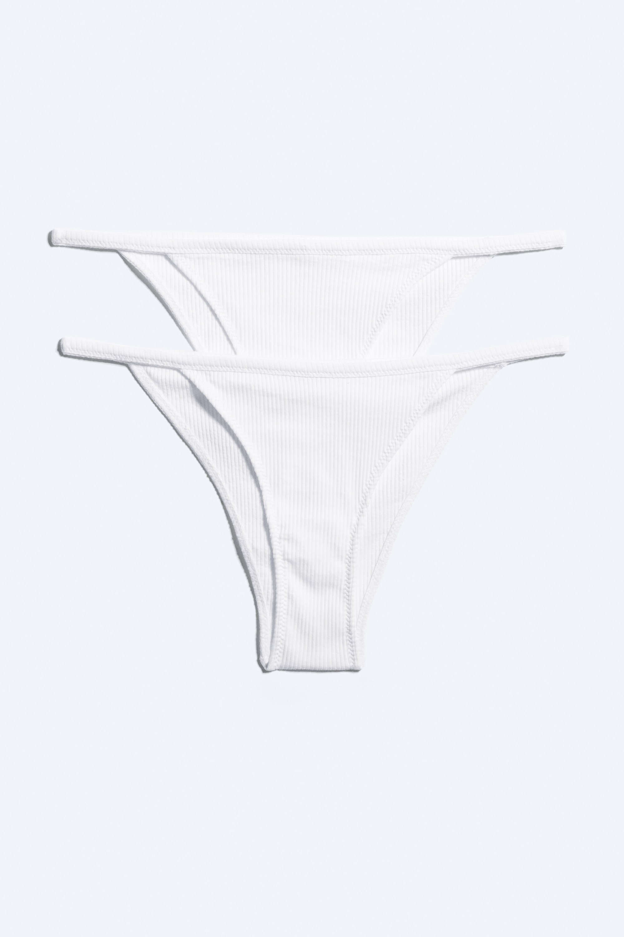 Le modÃ¨le porte une culotte tanga cÃ´telÃ©e blanche, avec une coupe taille basse et de fines brides latÃ©rales. La culotte est assortie Ã  un bralette cÃ´telÃ© blanc coordonnÃ©.