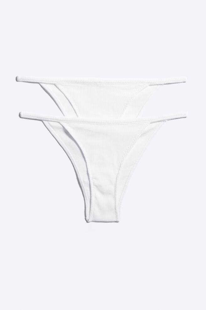 Um conjunto de duas cuecas tanga brancas em malha canelada minimalista estÃ¡ disposto na horizontal, exibindo o design frontal das peÃ§as empilhadas ligeiramente desalinhadas. Ambas as cuecas apresentam uma textura fina de malha canelada vertical, aberturas de perna de corte alto e finas alÃ§as laterais minimalistas. - 1