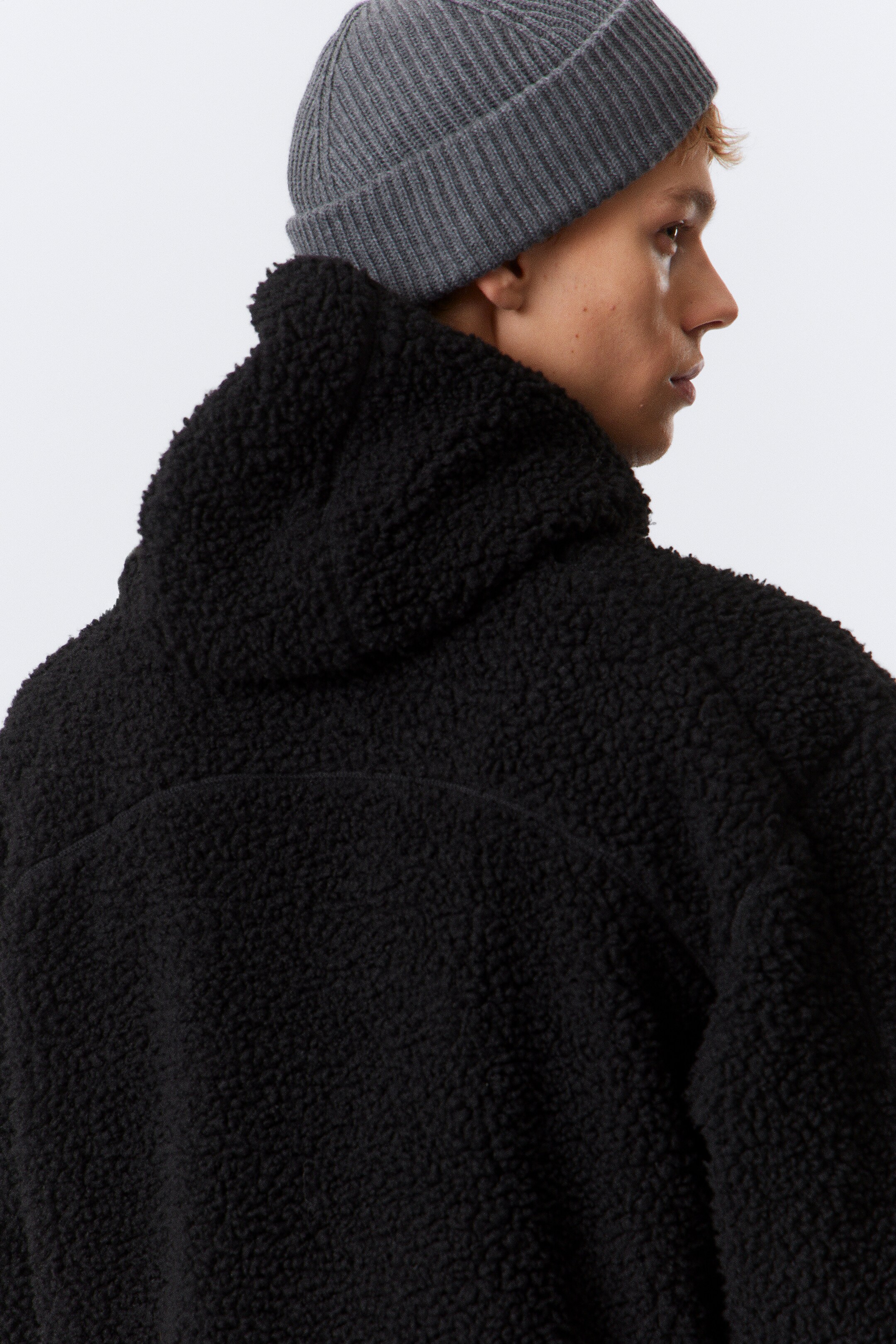 Zobrazit větší obrázek: Černá bunda z materiálu sherpa fleece je prezentována z pohledu zezadu a z boku, ukazuje svou texturovanou, kudrnatou látku a viditelnou kapuci. Také je nošena šedá čepice z vroubkovaného úpletu s ohrnutým lemem.