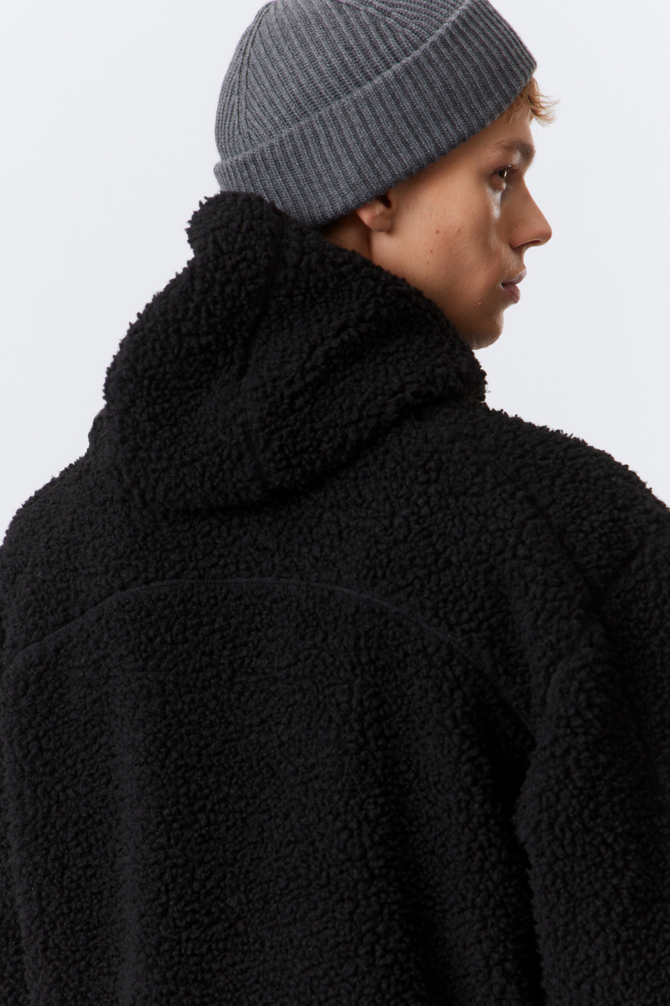 Visualizza immagine più grande: Una giacca in pile sherpa nero viene presentata da dietro e di lato, mostrando il suo materiale strutturato e riccioluto e un cappuccio visibile. È indossato anche un berretto a coste grigio con risvolto.
