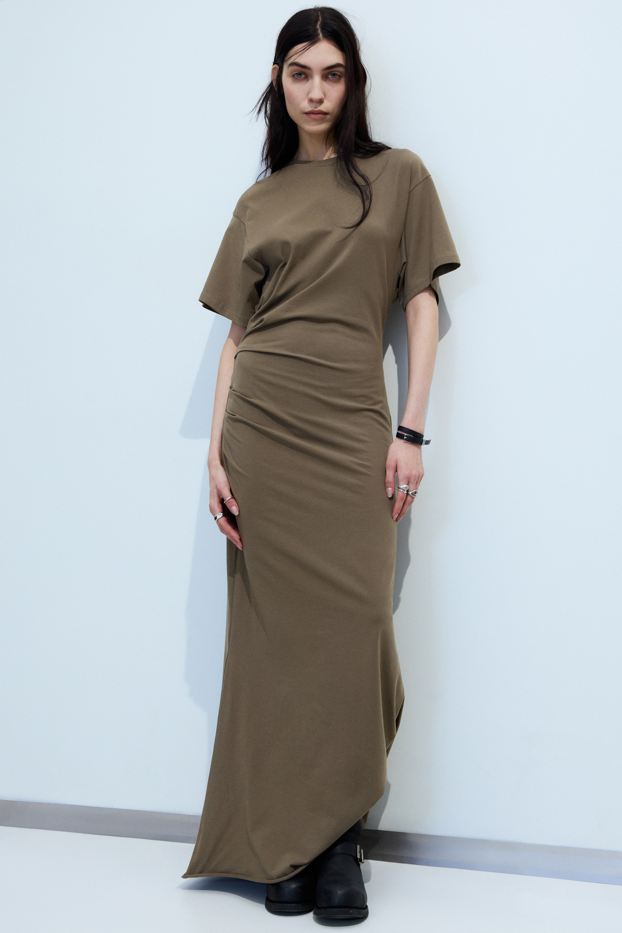 Se større billede: Den olivengrønne lange jerseykjole har en løs t-shirt stil top med korte ærmer og en rund halsudskæring, der går over i en tætsiddende midtersektion med rynker på venstre side, og flyder ud i et langt skørt, der draperer ned til gulvet.