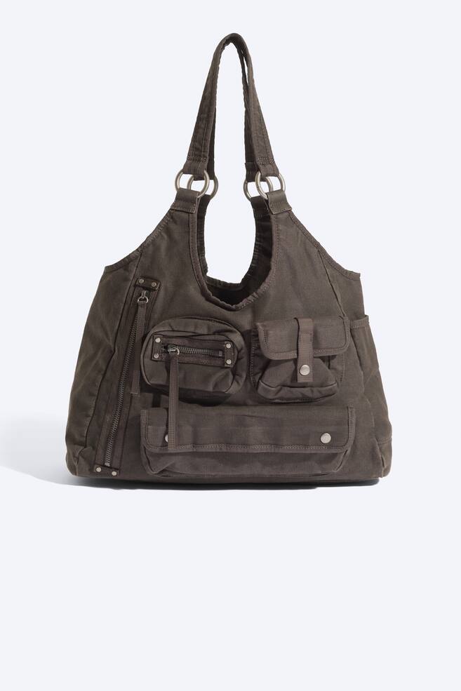 Eine dunkelbraune Canvas-UmhÃ¤ngetasche ist von vorne zu sehen und prÃ¤sentiert eine lÃ¤ssig fallende Silhouette sowie mehrere externe Utility-FÃ¤cher. Die Tasche verfÃ¼gt Ã¼ber zwei Schulterriemen, die mit silberfarbenen Metallringen befestigt sind, verschiedene ReiÃverschluss- und KlappenfÃ¤cher mit Druckverschluss auf der Vorderseite sowie seitliche SteckfÃ¤cher. - 1