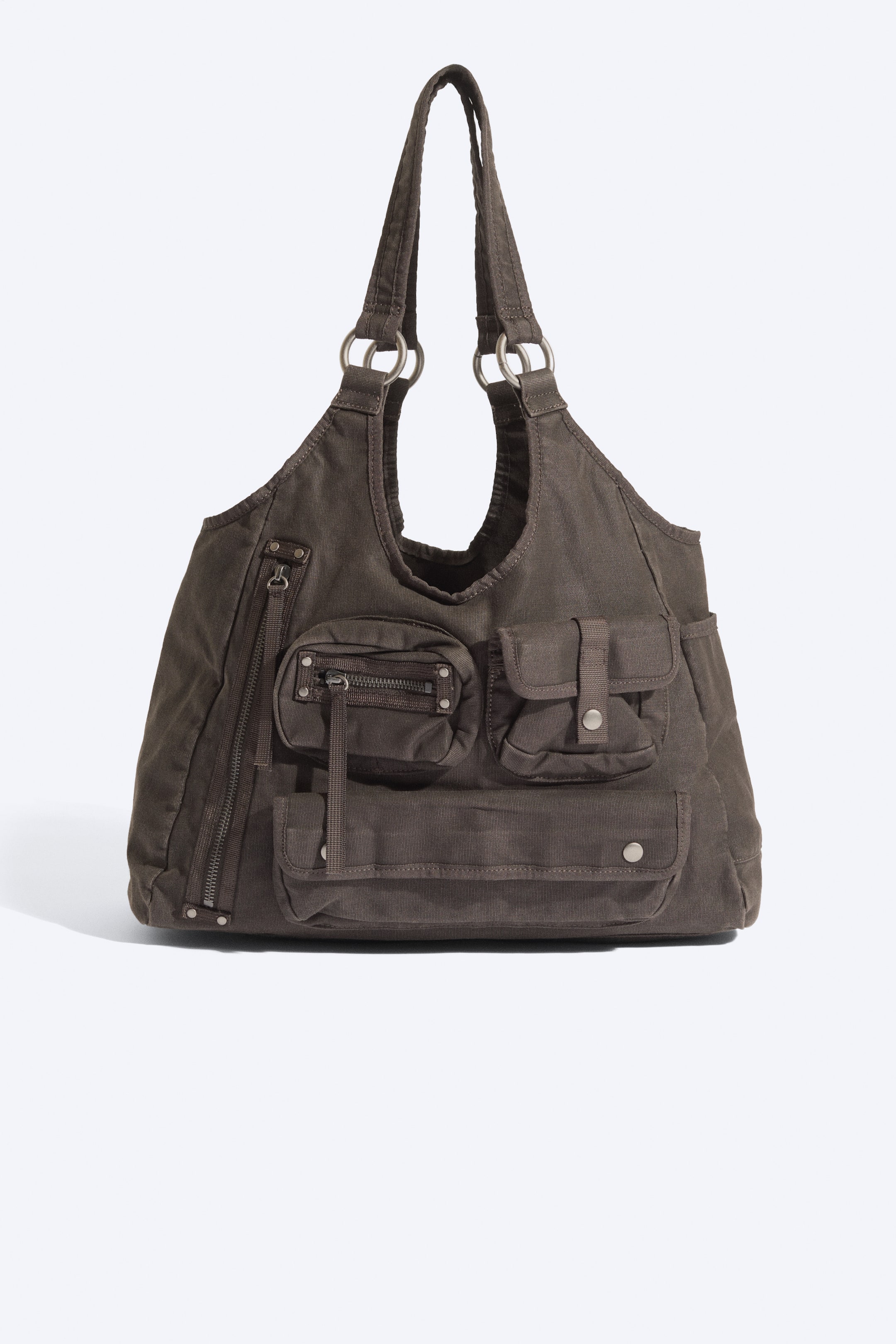 Größeres Bild anzeigen: Eine dunkelbraune Canvas-Schultertasche ist von vorne zu sehen und zeigt eine lässig fallende Silhouette sowie mehrere äußere Utility-Fächer. Die Tasche verfügt über zwei Schulterriemen, die mit silberfarbenen Metallringen befestigt sind, verschiedene Fronttaschen mit Reißverschluss und Druckknopfklappe sowie seitliche Einschubtaschen.