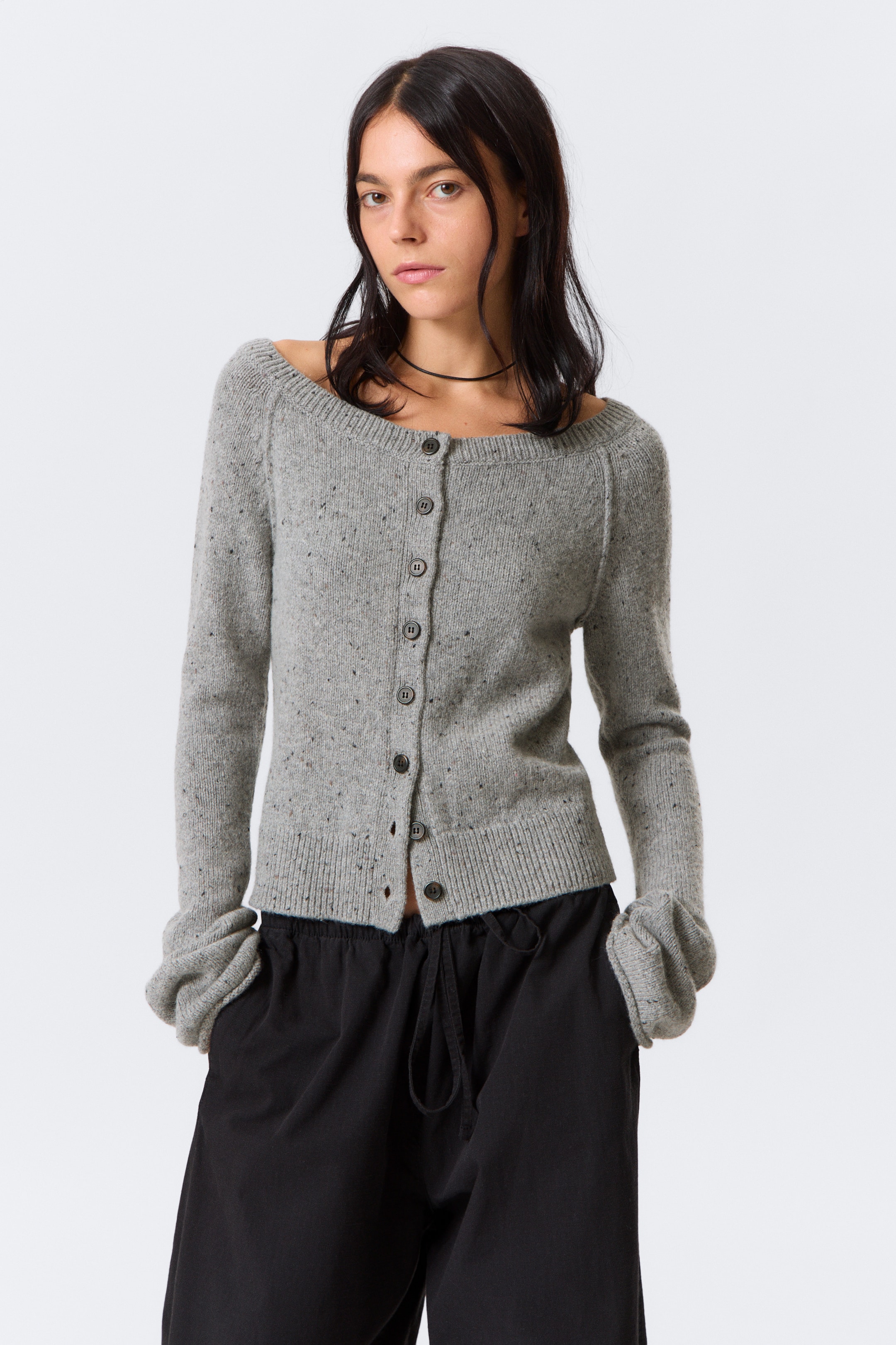 Agrandir l'image: Un cardigan boutonné gris clair avec un col large est porté par le mannequin. Ses manches trompette se rassemblent autour de ses mains alors qu'elle les glisse dans les poches de son pantalon, et les boutons du bas sont défaits.