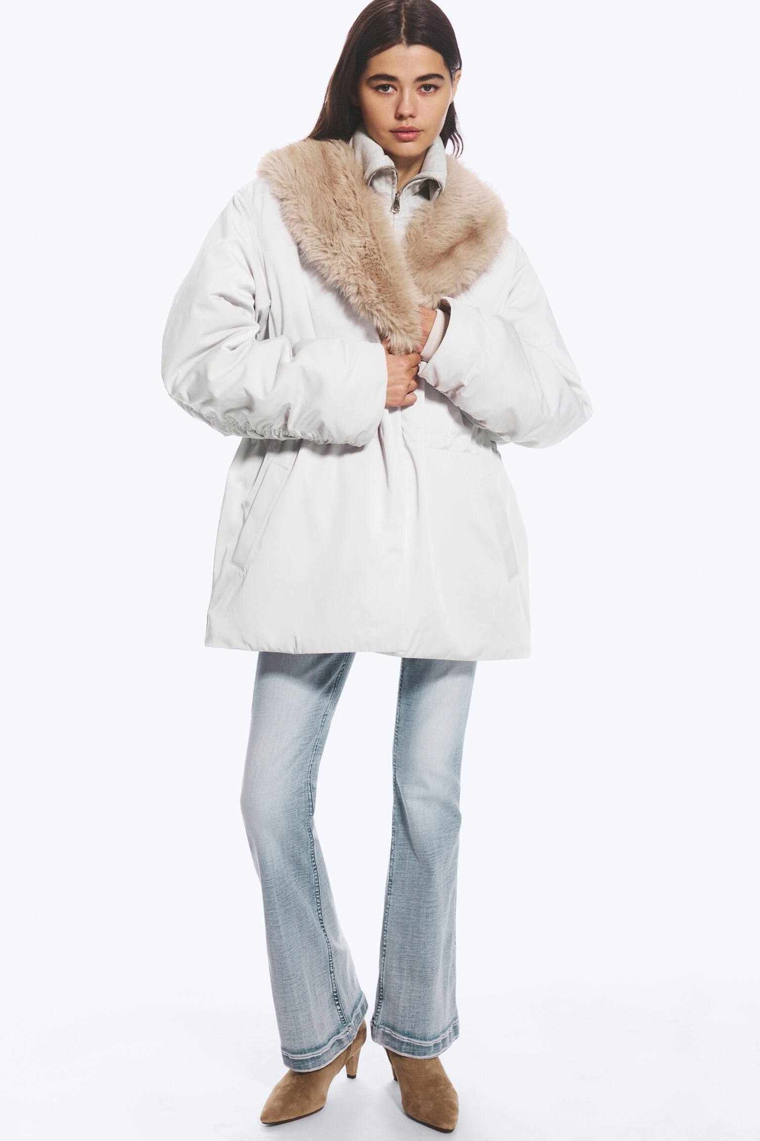 A light grey padded jacket featuring a beige faux fur shawl collar and a cinched waist is being worn by the model, who is holding the front of the garment. The jacket is layered over a grey zip-up top and paired with light-wash flared jeans. - 1