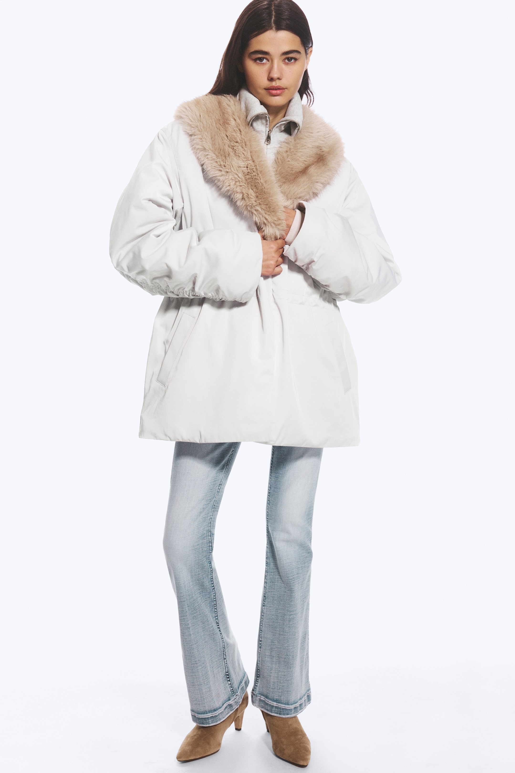 View larger image: A light grey padded jacket featuring a beige faux fur shawl collar and a cinched waist is being worn by the model, who is holding the front of the garment. The jacket is layered over a grey zip-up top and paired with light-wash flared jeans.