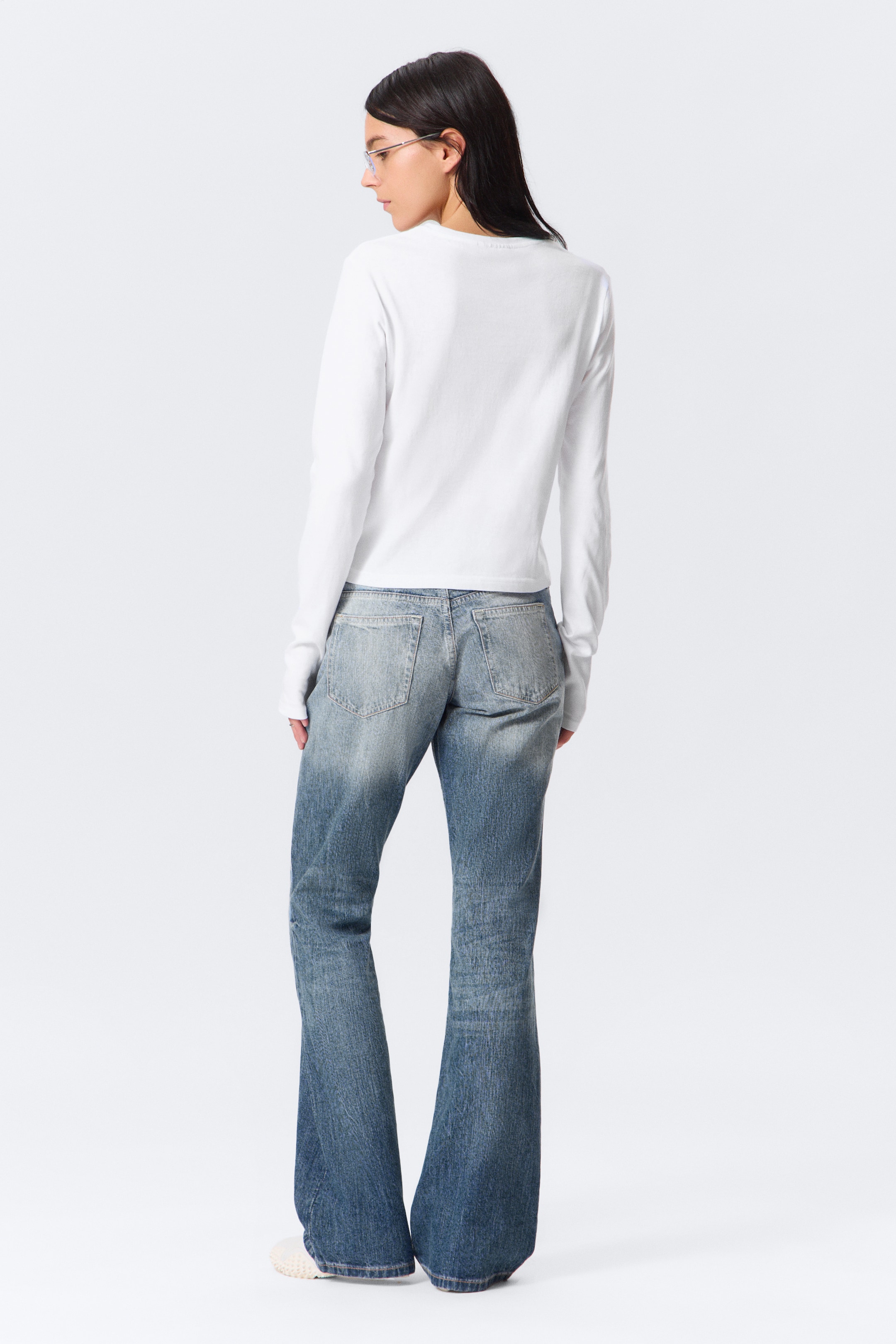 Se større billede: En hvid langærmet top bæres sammen med et par blå distressed jeans, præsenteret bagfra. Jeansene har en bootcut eller flare ben og en falmet vask, mens toppen fremviser en slank pasform og en lige kant.