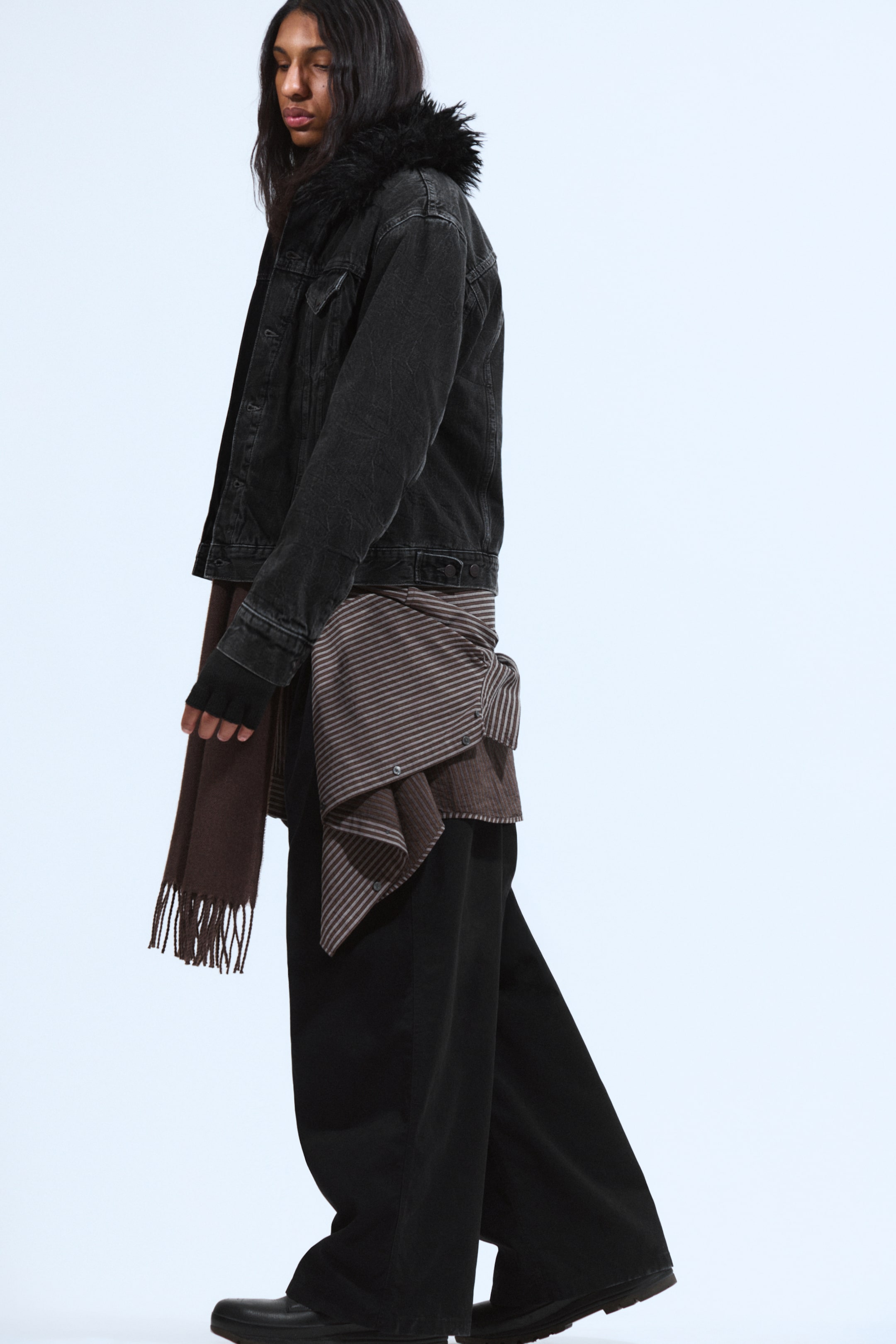 View larger image: A black distressed denim jacket, featuring a black faux fur collar, is being presented from a side angle. Black wide-leg pants are being worn, with a brown striped shirt being tied around the waist and a brown fringed scarf draping from the left hand.