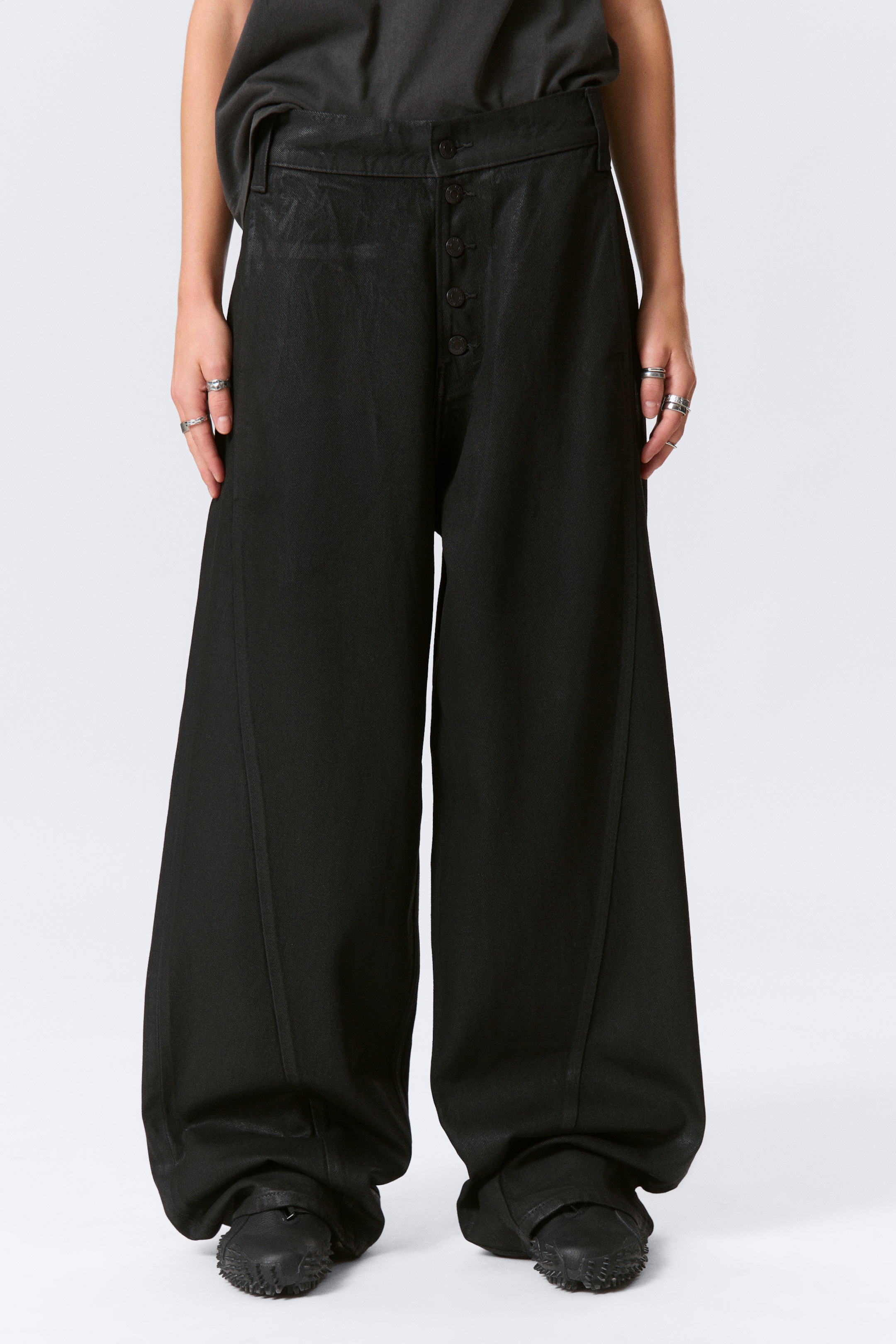 View larger image: A pair of black rigid loose-fit jeans with a baggy leg is being worn by the model. They feature an exposed button fly and prominent twin-needle seams along the legs, with the fabric pooling over the model's black textured shoes.