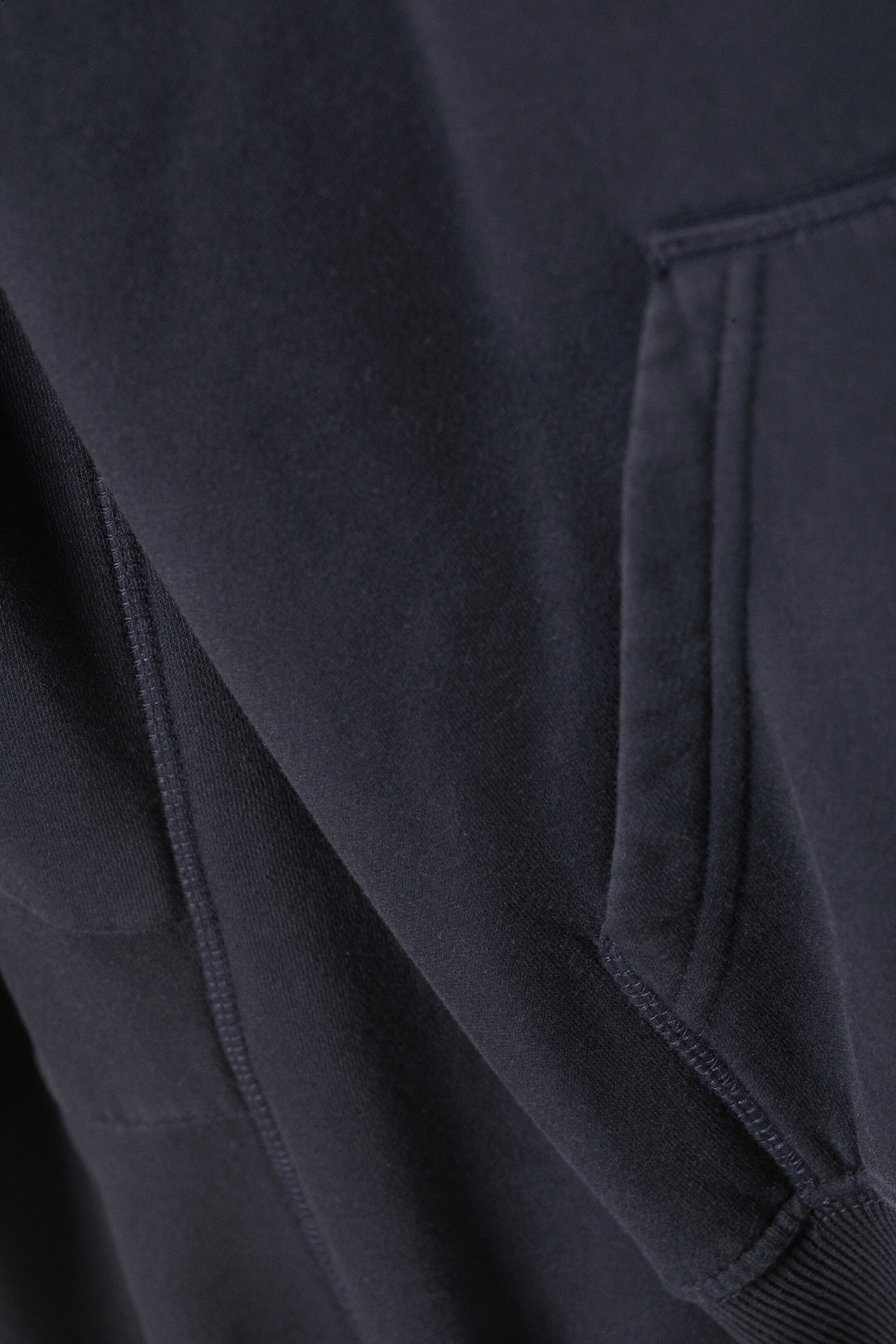Größeres Bild ansehen: Ein dunkel gefärbter Hoodie wird in einer Nahaufnahme von vorne gezeigt. Er besteht aus einem weichen, strukturierten Stoff und verfügt über eine auffällige Kängurutasche mit sichtbaren Nähten. Ein breiter gerippter Saum ist ebenfalls am unteren Rand sichtbar.