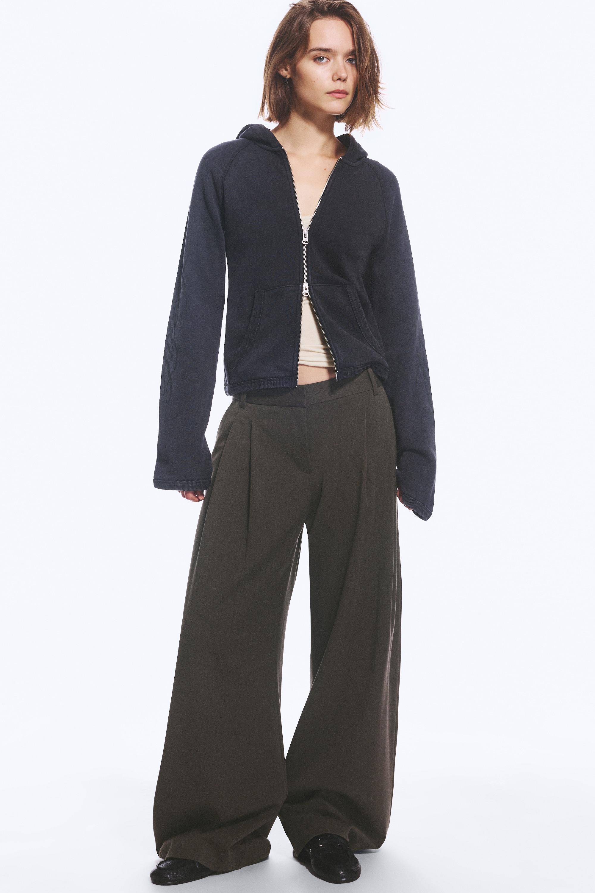 Donkergrijze geplooide pantalons met lage taille worden frontaal getoond, met een wijde pijpen-silhouet en diepe plooien aan de voorkant. De broek heeft een gestructureerde tailleband met riemlussen en subtiele steekzakken aan de zijkant.