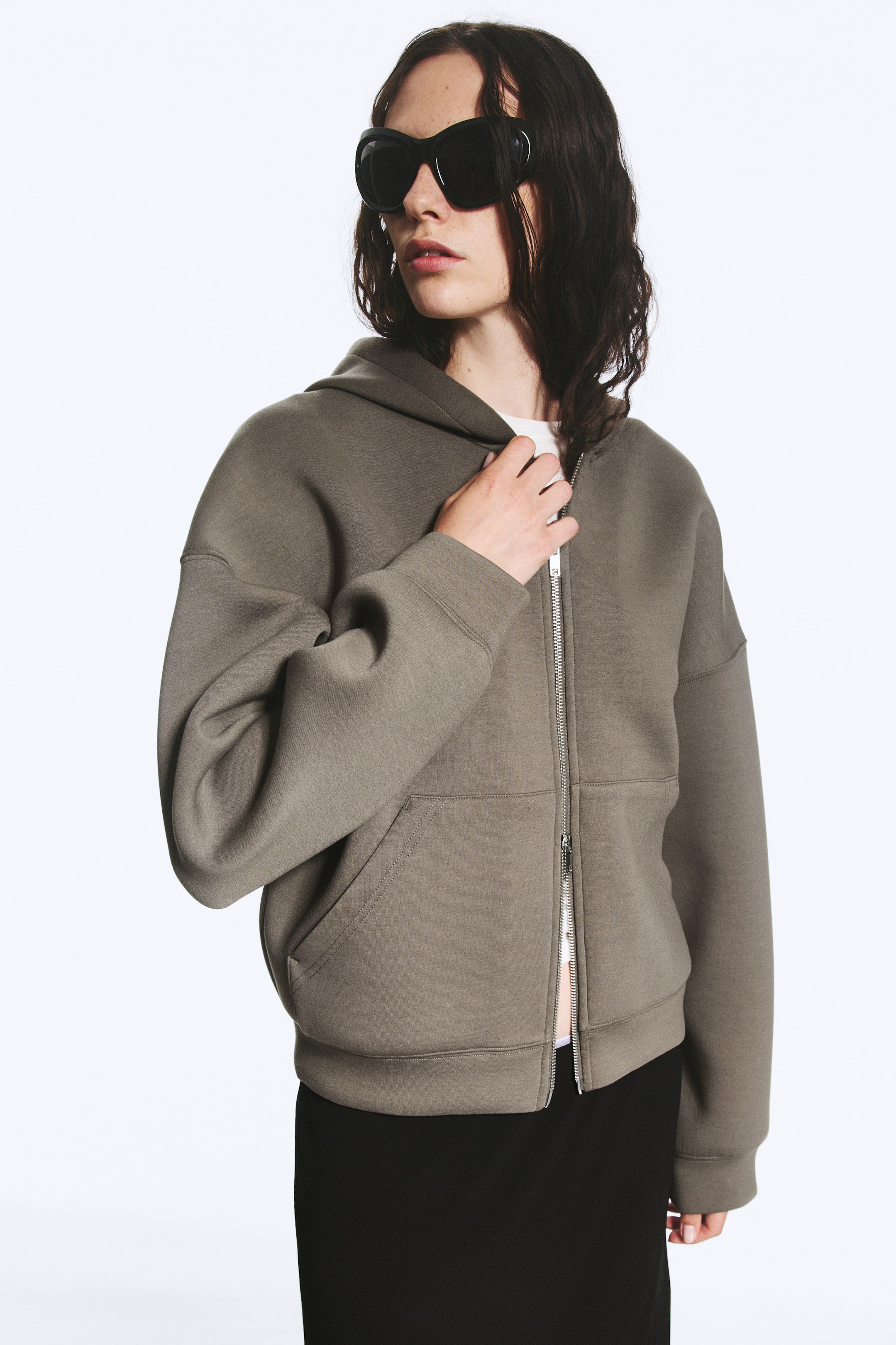 Agrandir l'image: Un hoodie à fermeture éclair complète de couleur kaki-brun avec une coupe décontractée et un tissu en maille lisse est présenté, mettant en avant une capuche, une fermeture éclair en métal de longueur complète partiellement ouverte depuis le bas pour révéler un t-shirt blanc, ainsi que des poches kangourou à l'avant.