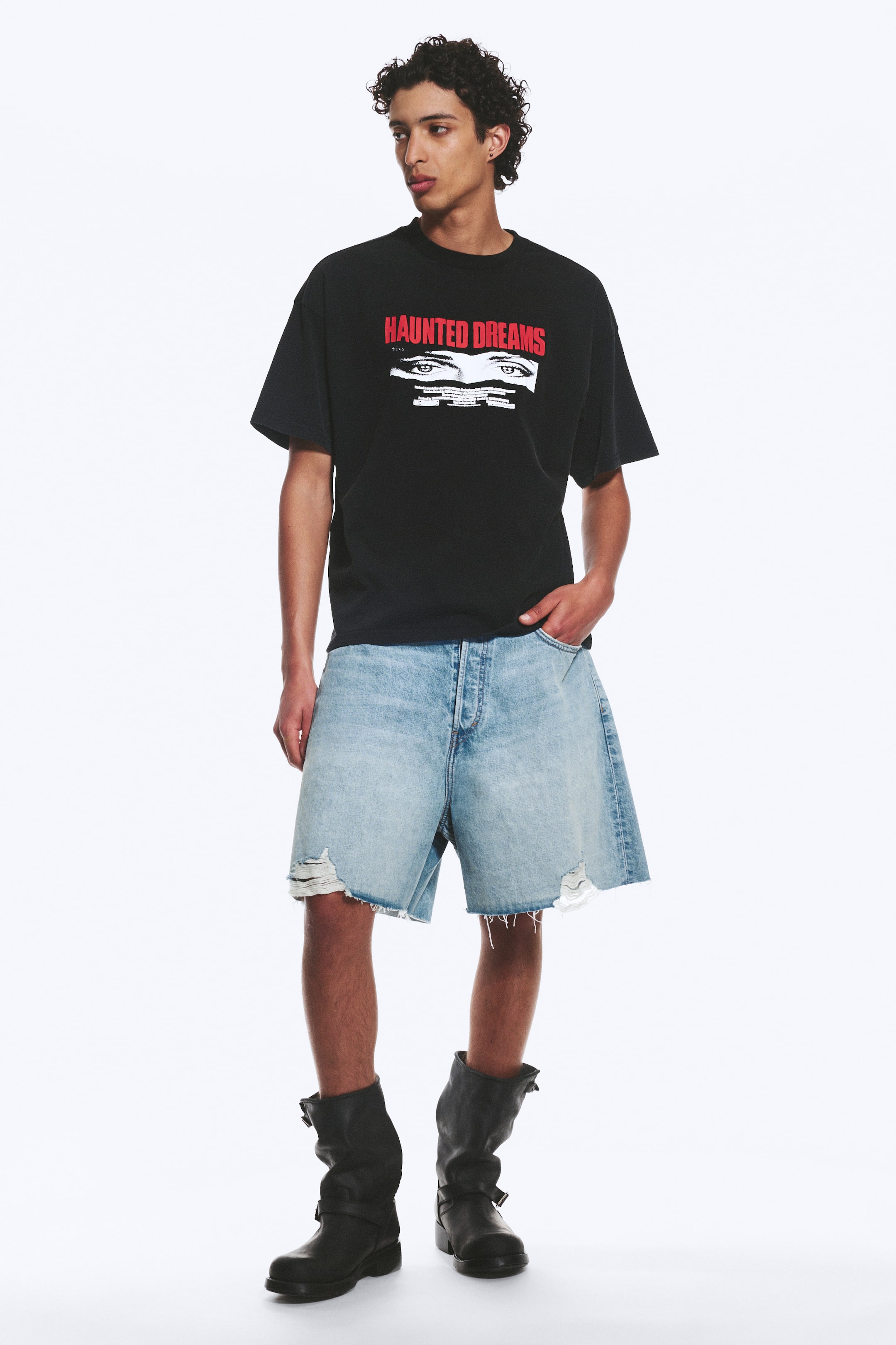 Grotere afbeelding bekijken: Een zwarte oversized t-shirt, met een rode "HAUNTED DREAMS" grafische print boven witte en zwarte ogen, wordt gedragen met lichtgewassen distressed denim shorts met rafelige zomen en een zichtbare knoopsluiting, en zwarte laarzen tot halverwege de kuit met gespsluitingen.