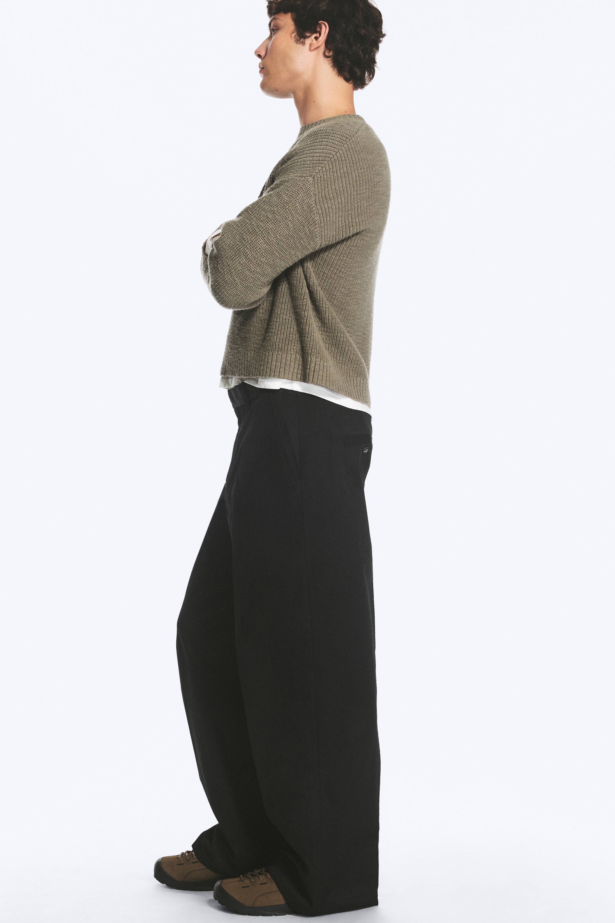 Vezi imaginea mai mare: Un model poartă o pereche de pantaloni chino negri, cu croială lejeră, fiind surprins din profil. Pantalonii au o siluetă cu picior larg și sunt asortați cu un pulover scurt, din tricot ribat, de culoare verde-oliv, și pantofi maro.