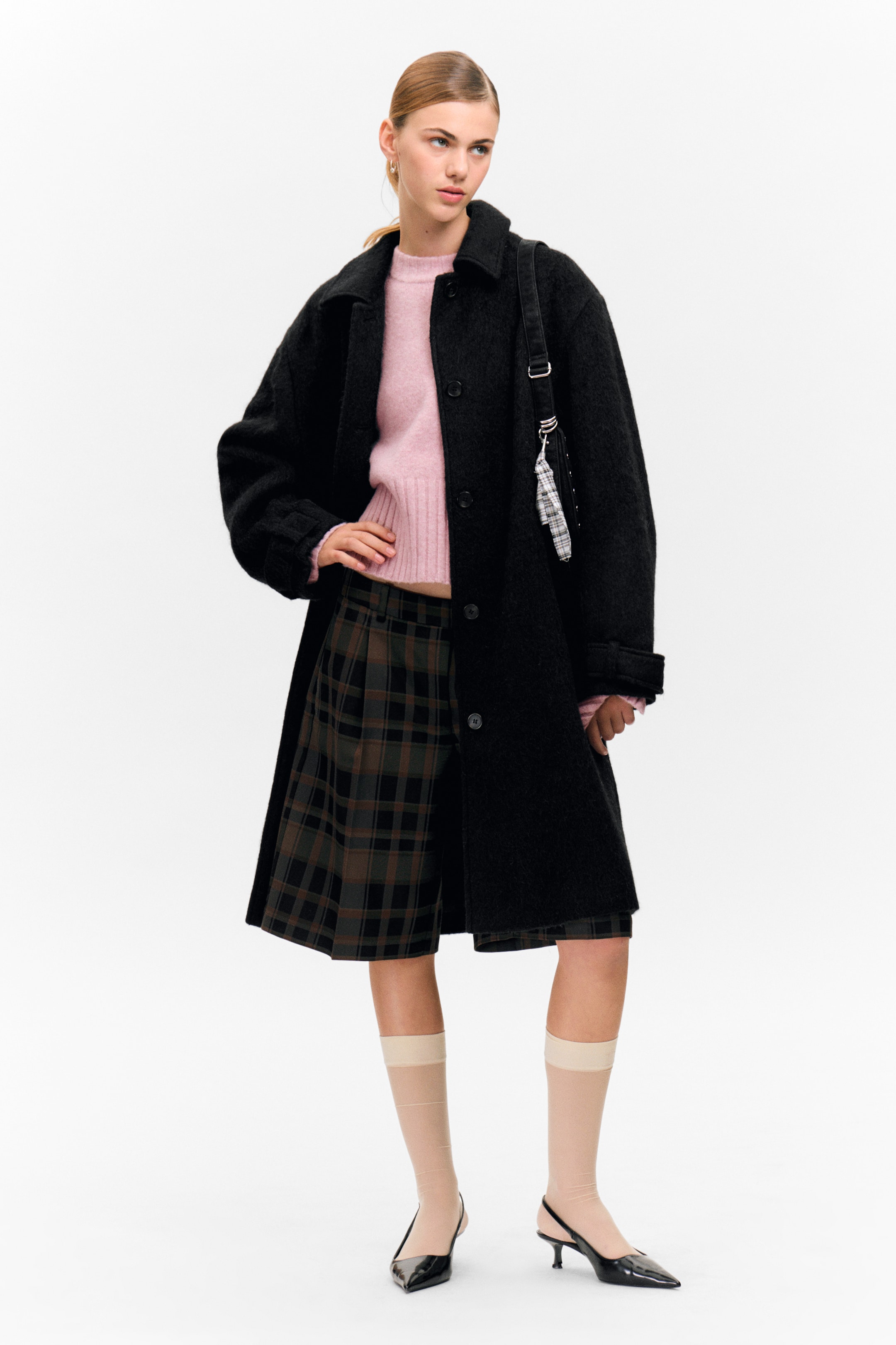 View larger image: A front-facing long, black textured coat with a classic collar and visible front buttons is being worn over a light pink ribbed knit crewneck sweater. Dark green and brown plaid pleated shorts are visible beneath the coat, along with sheer beige knee-high socks and black pointed-toe slingback kitten heels.