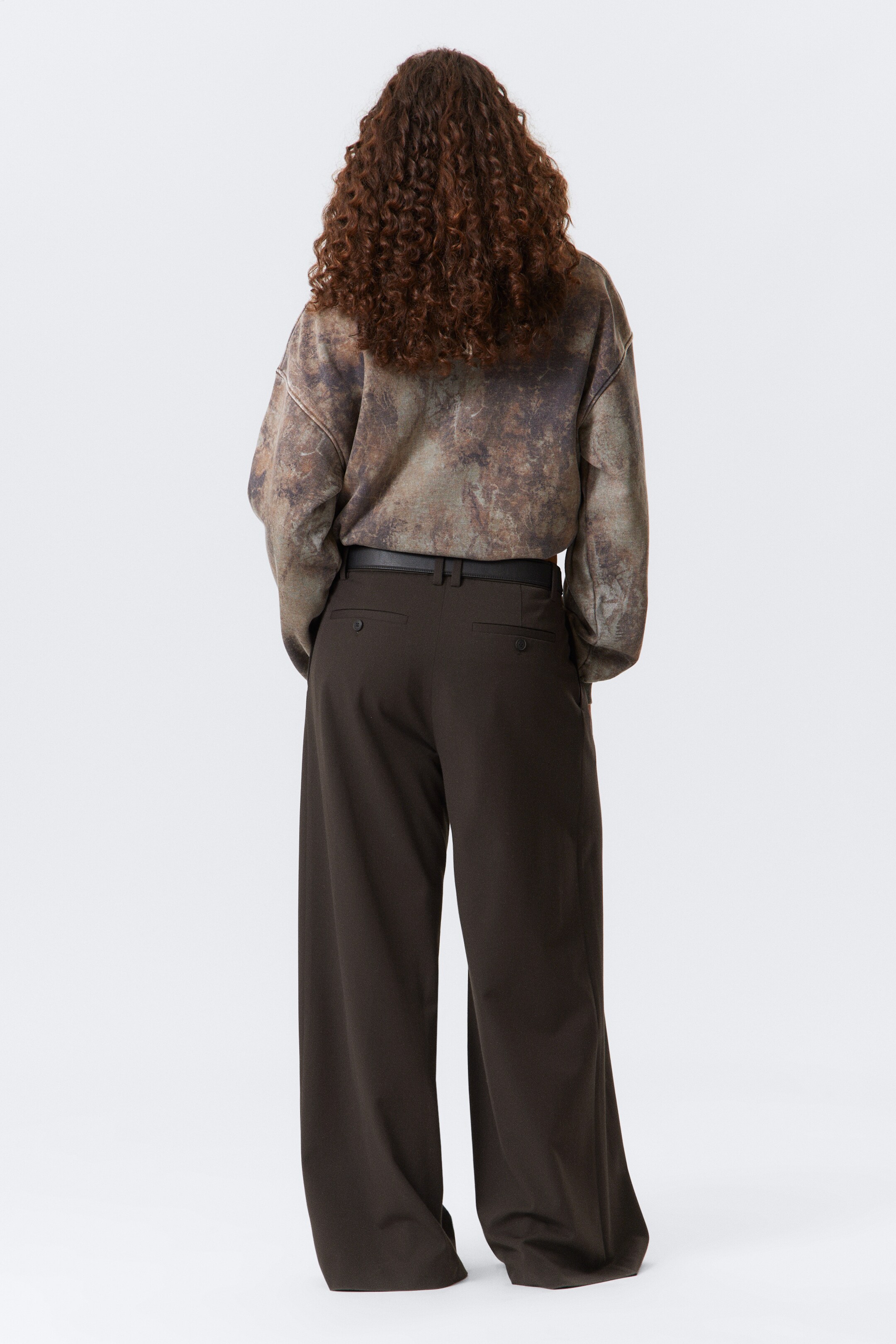 Visualizza immagine più grande: Un top a maniche lunghe con un motivo screziato in tonalità terrose è indossato, infilato in pantaloni a gamba larga di colore marrone scuro. I pantaloni presentano passanti per cintura, una cintura nera e due tasche posteriori a filetto con chiusure a bottone.