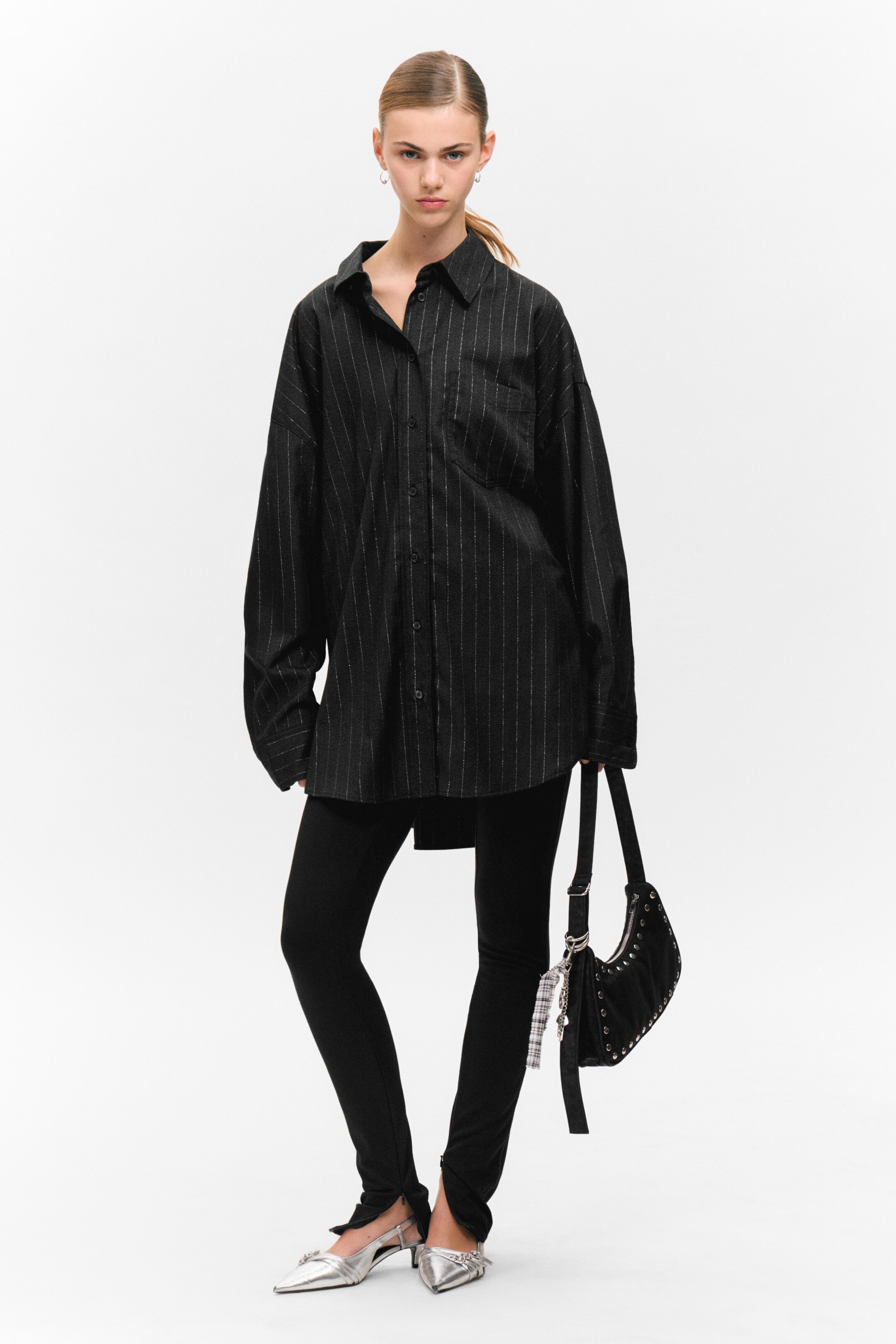 Agrandir l'image: Une chemise oversize à rayures noires est présentée, avec un col pointu, des manches longues et une poche poitrine. Des leggings noirs à étriers sont portés en dessous, avec des chaussures slingback à bout pointu argentées, accentuées par des ornements, portées aux pieds. Un sac à main noir en forme de croissant avec des clous et un charme à carreaux est tenu.