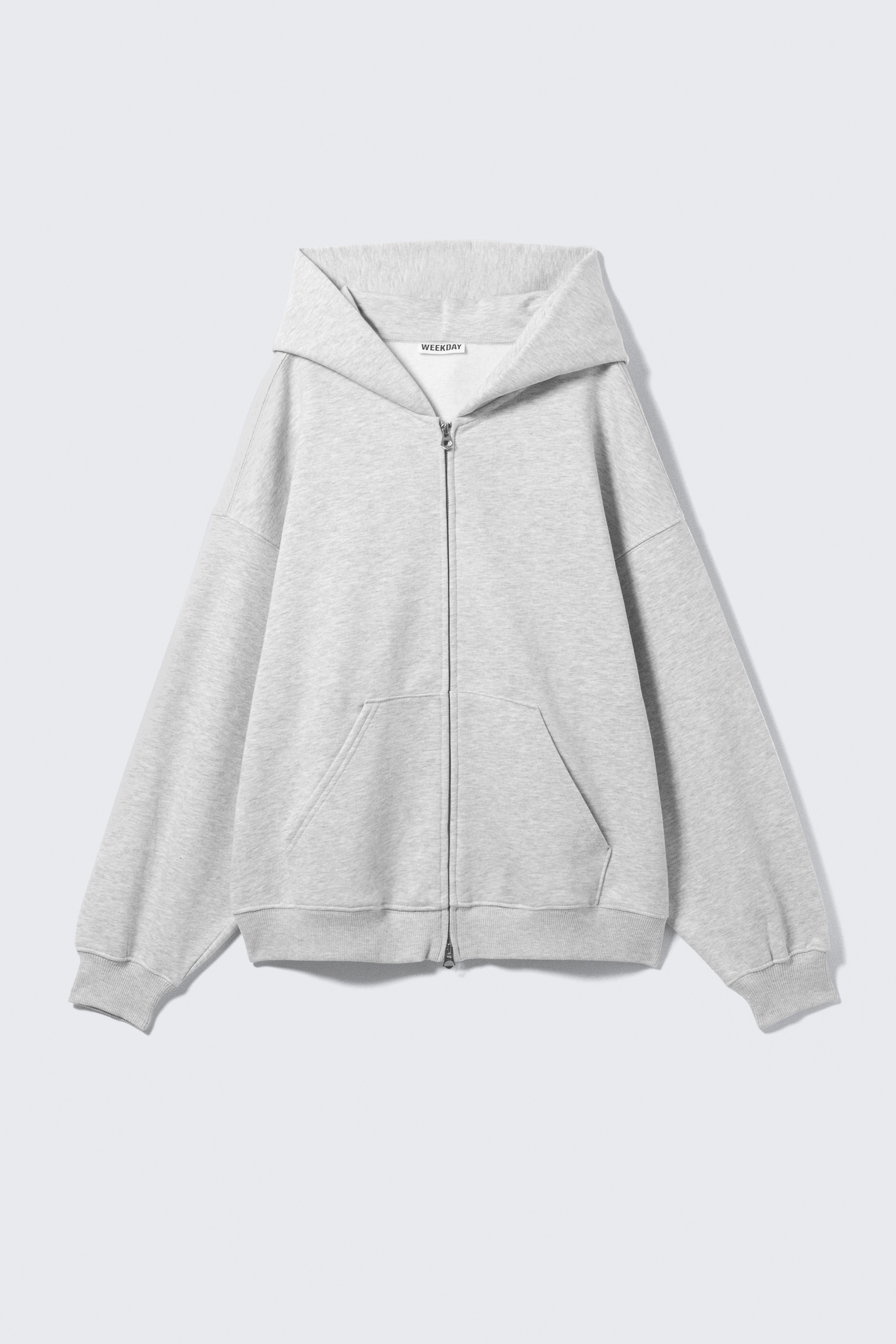 Ingrandisci l'immagine: Una felpa con cappuccio grigia chiara e pesante con zip è mostrata dalla parte anteriore, con una silhouette oversize e rilassata in un morbido tessuto felpato misto cotone. Include un ampio cappuccio a doppio strato senza laccetti, spalle scese, una zip argentata visibile a doppio verso, grandi tasche frontali e polsini e orlo a coste.