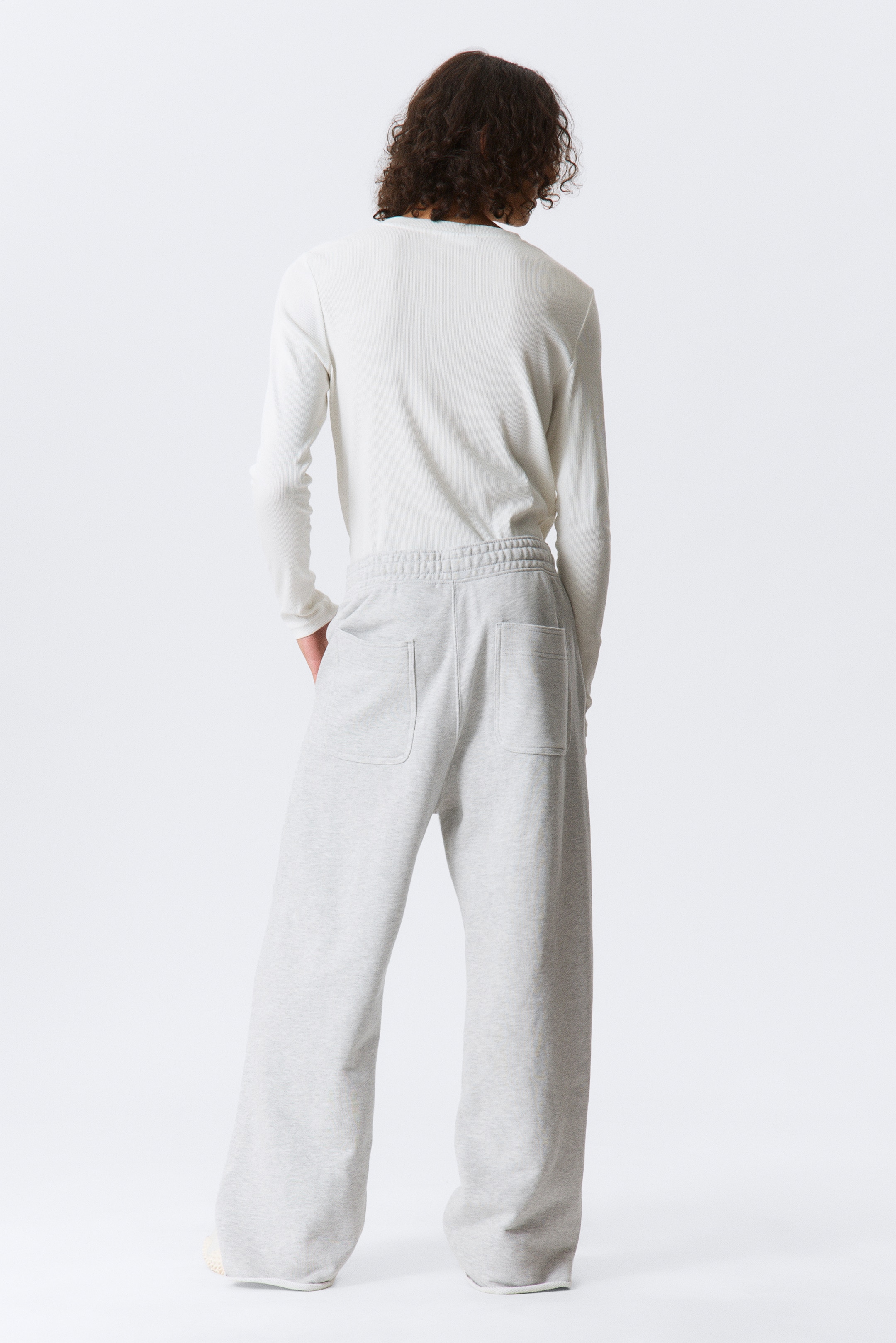 Grotere afbeelding bekijken: Een wit shirt met lange mouwen wordt gedragen met lichtgrijze joggingbroek, getoond van de achterkant. Het shirt heeft een ronde hals en lange mouwen, terwijl de wijde joggingbroek een elastische tailleband en twee opgestikte zakken aan de achterkant heeft.