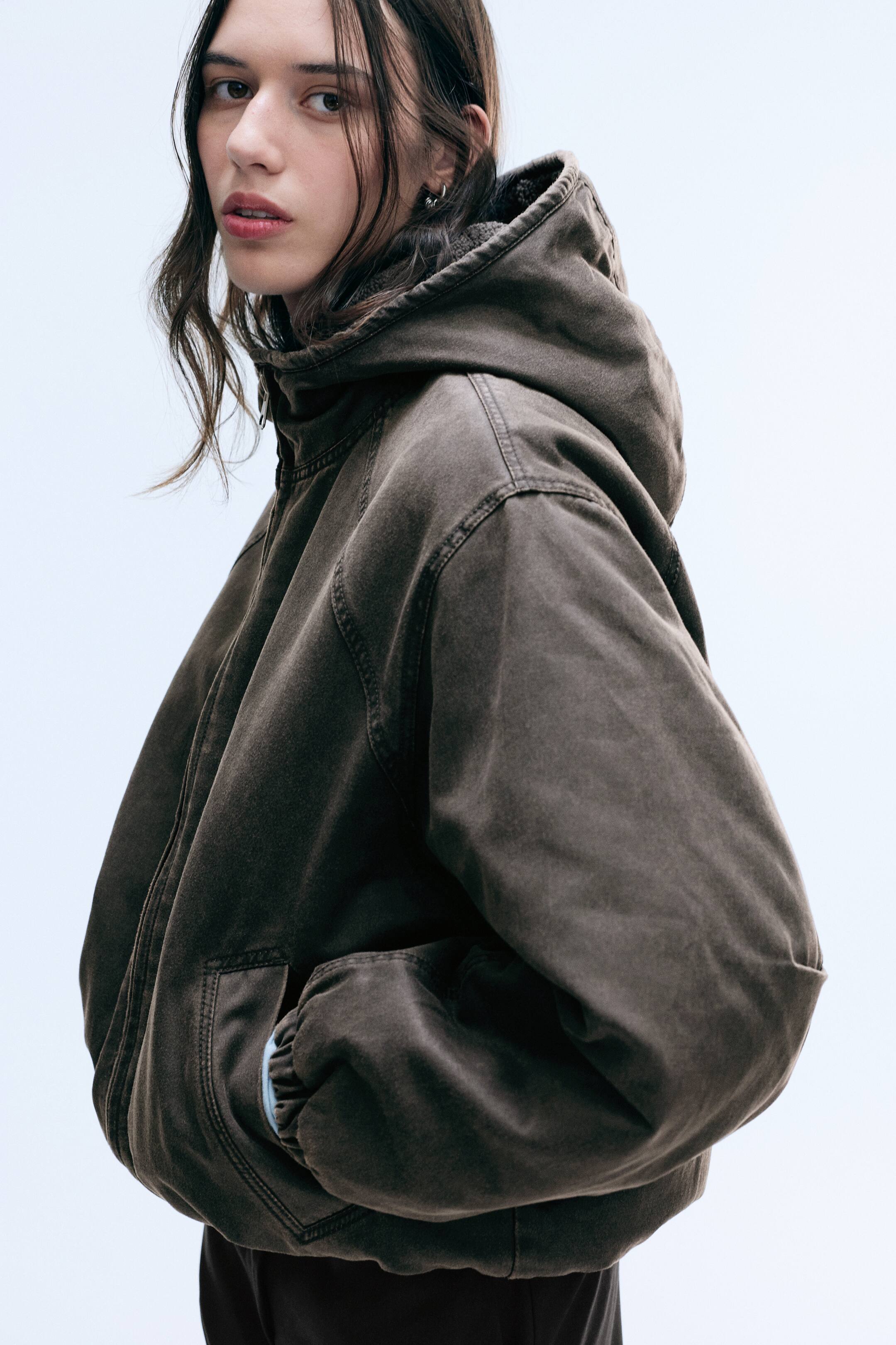 View larger image: A brown, washed canvas hooded jacket is being worn by the model, who is standing in profile with one hand resting in the side pocket. The jacket features a front zip fastening, seam detailing along the shoulders, and elasticated cuffs.