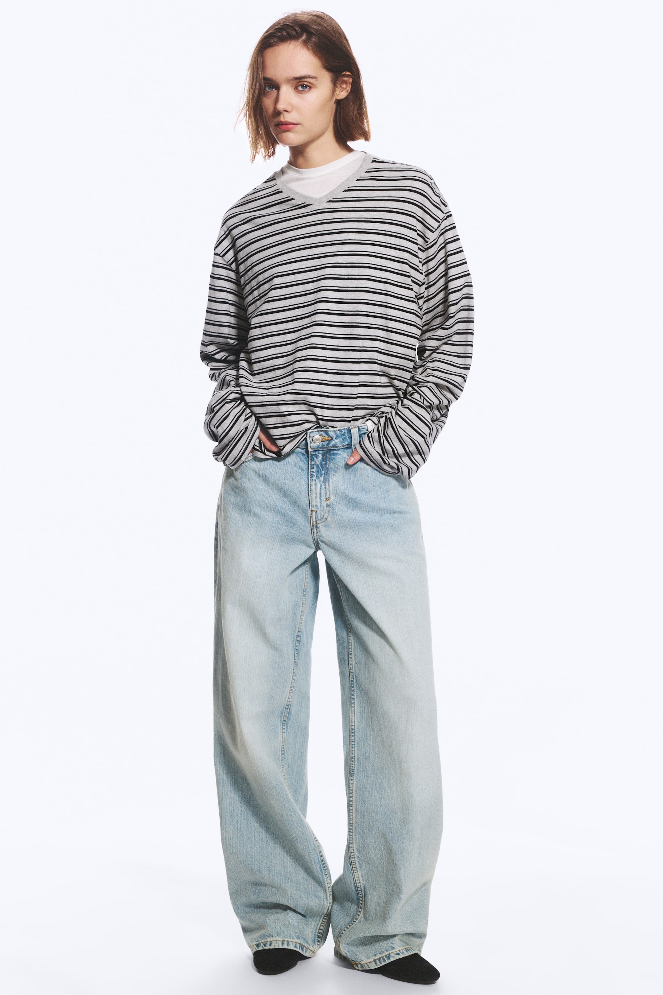 Agrandir l'image: Une paire de jeans délavés clair, taille basse, à la coupe ample, aux jambes larges et à la longueur longue, est portée par le mannequin. Le mannequin se tient debout, les mains posées dans les poches avant du jean, associé à une chemise à manches longues rayée gris et noir.