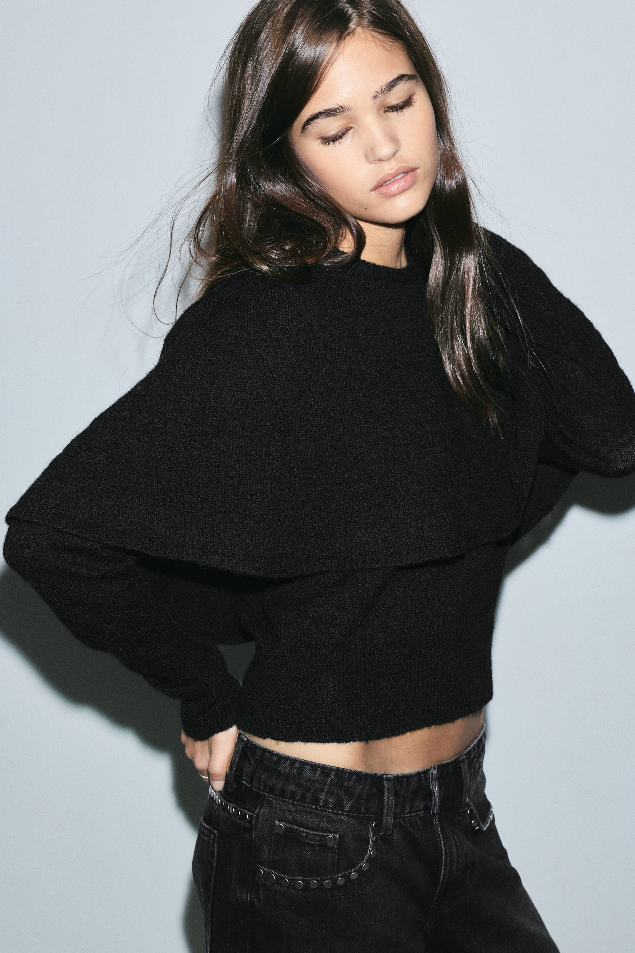 Größeres Bild anzeigen: Ein schwarzer Strickpullover mit Rundhalsausschnitt, langen Ärmeln und einem drapierten Cape-Overlay über den Schultern wird vom Model getragen. Das figurbetonte Oberteil ist mit Rippbündchen versehen und mit dunkel gewaschenen Denim-Jeans kombiniert.