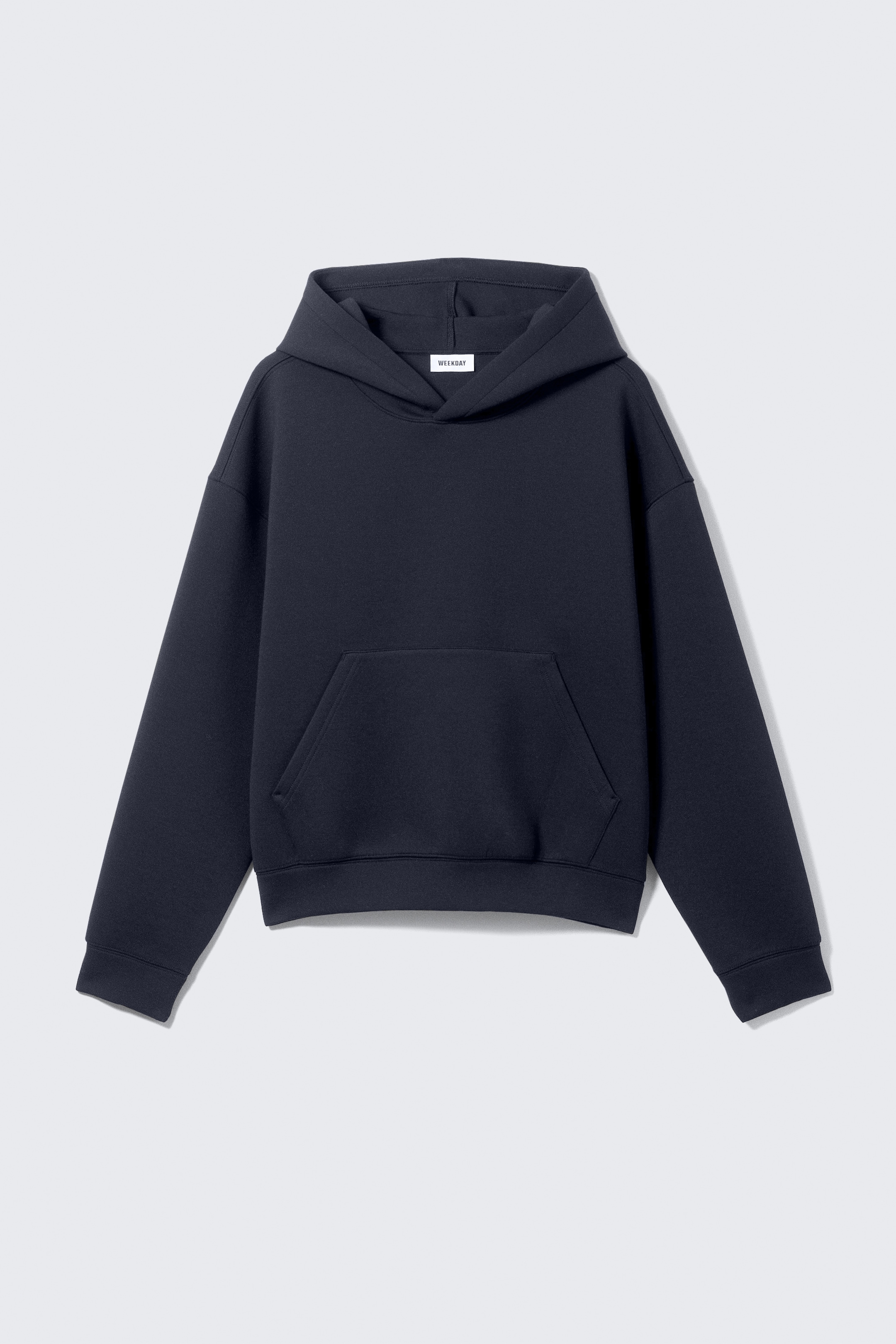 Größeres Bild anzeigen: Ein dunkelblauer Hoodie liegt flach da, von vorne präsentiert, und zeigt eine entspannte Passform mit überschnittenen Schulternähten. Er verfügt über eine große Kapuze, eine geräumige Kängurutasche und breite Bündchen und Saum, mit dem Markenlabel 'WEEKDAY', das im Inneren der Kapuze sichtbar ist.