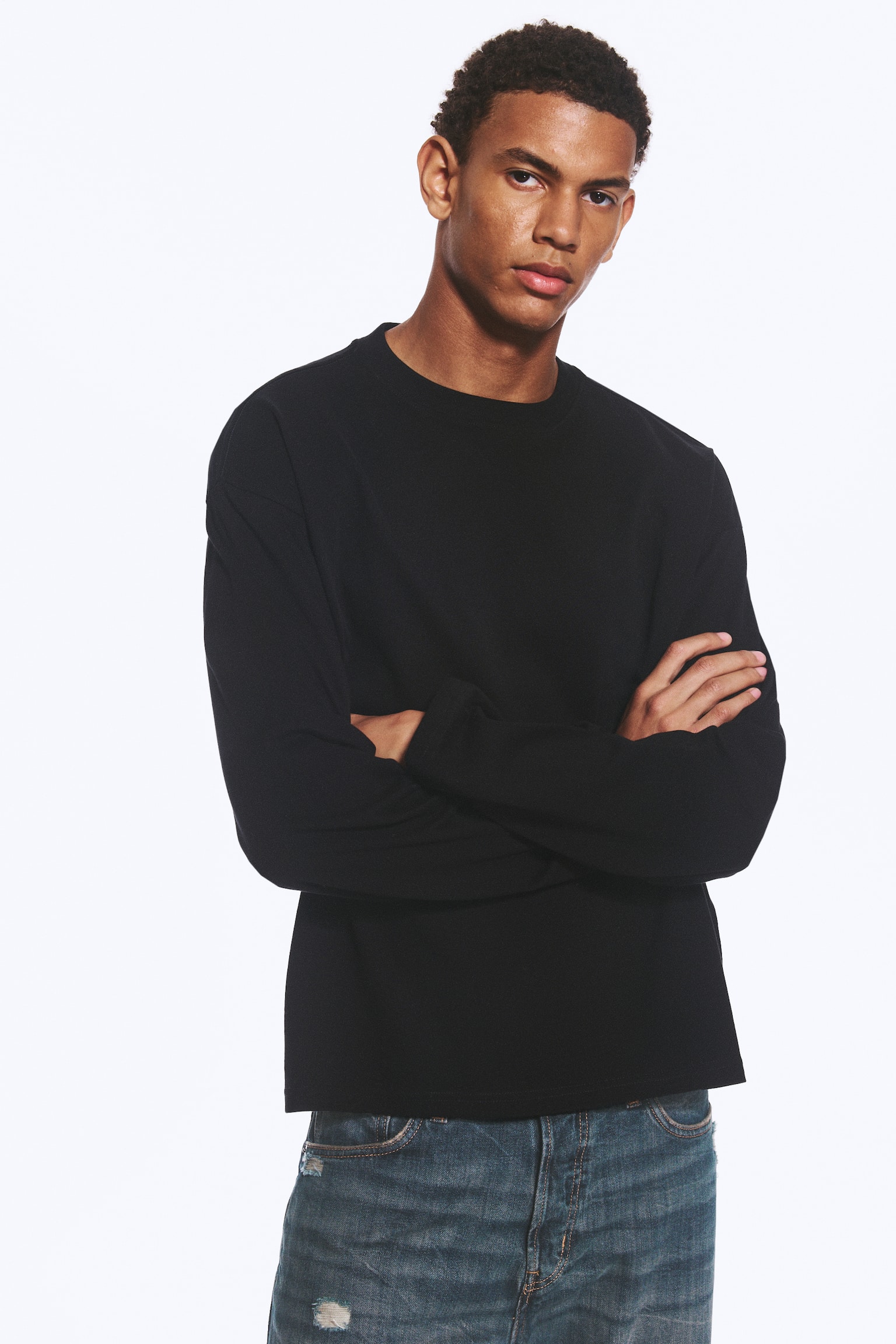 La modelo lleva una camiseta negra de manga larga, de corte recto y holgado, con cuello redondo y bajo recto. La modelo está de pie con los brazos cruzados, combinando la parte superior de ajuste holgado con unos vaqueros azules. - 1