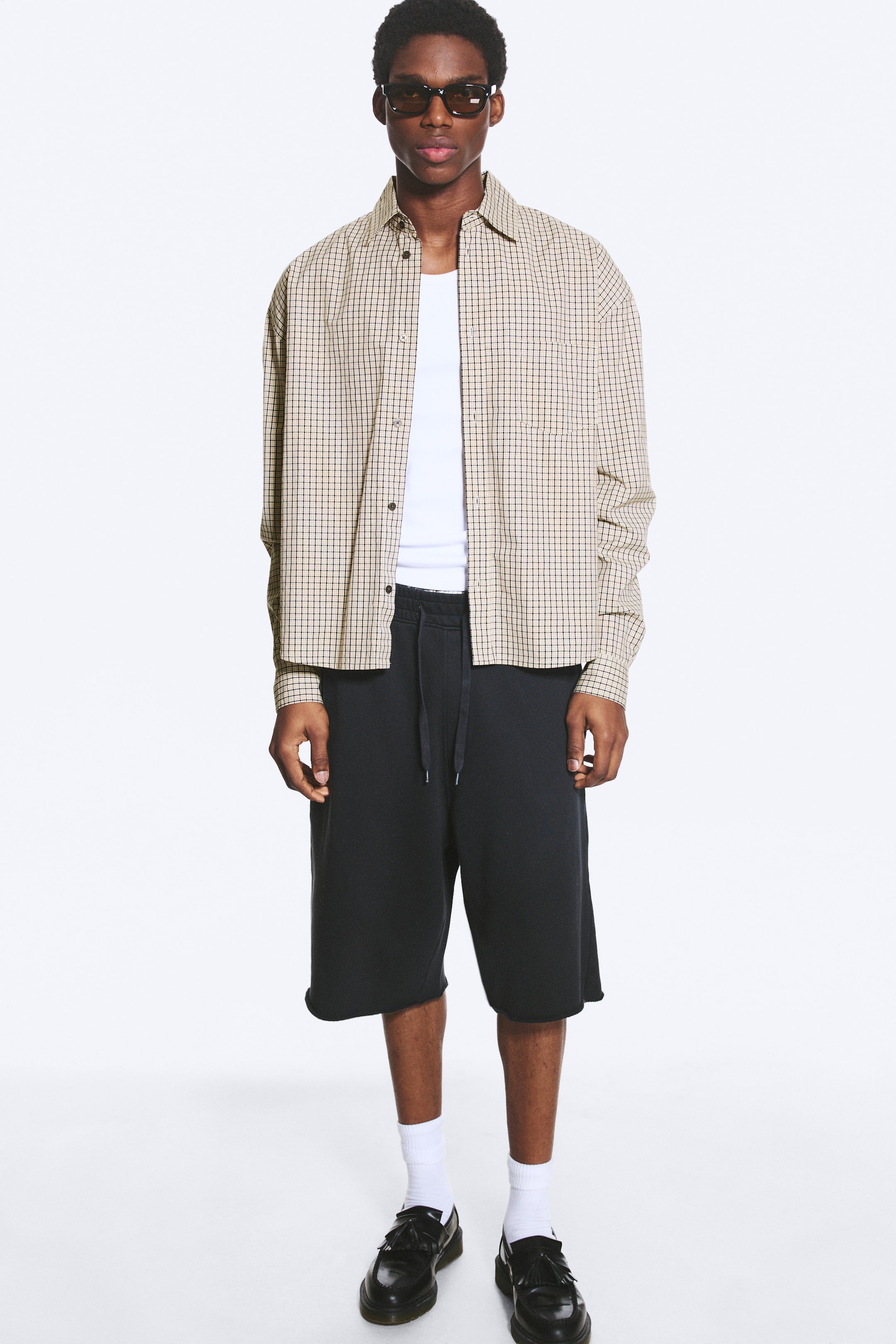 Agrandir l'image: Une chemise beige à carreaux boutonnée est superposée à un t-shirt blanc à col rond, associée à un short noir longueur genou avec une taille à cordon de serrage et des ourlets bruts. Ce look est complété par des chaussettes blanches montantes, des mocassins noirs à pampilles et des lunettes de soleil rectangulaires foncées.