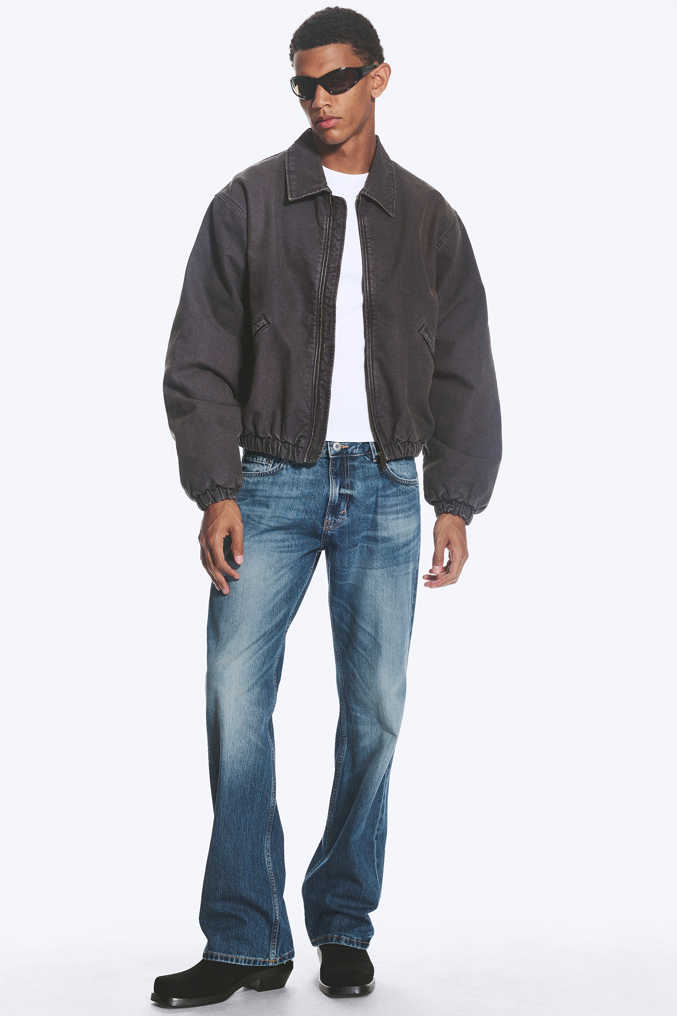 Agrandir l'image: Une veste gris foncé à col rabattu avec fermeture zippée sur le devant et poignets et ourlet élastiqués, offrant une coupe oversize sur un t-shirt blanc. Un jean bootcut bleu délavé présente un fini plissé et est porté avec des bottes noires à bout pointu.