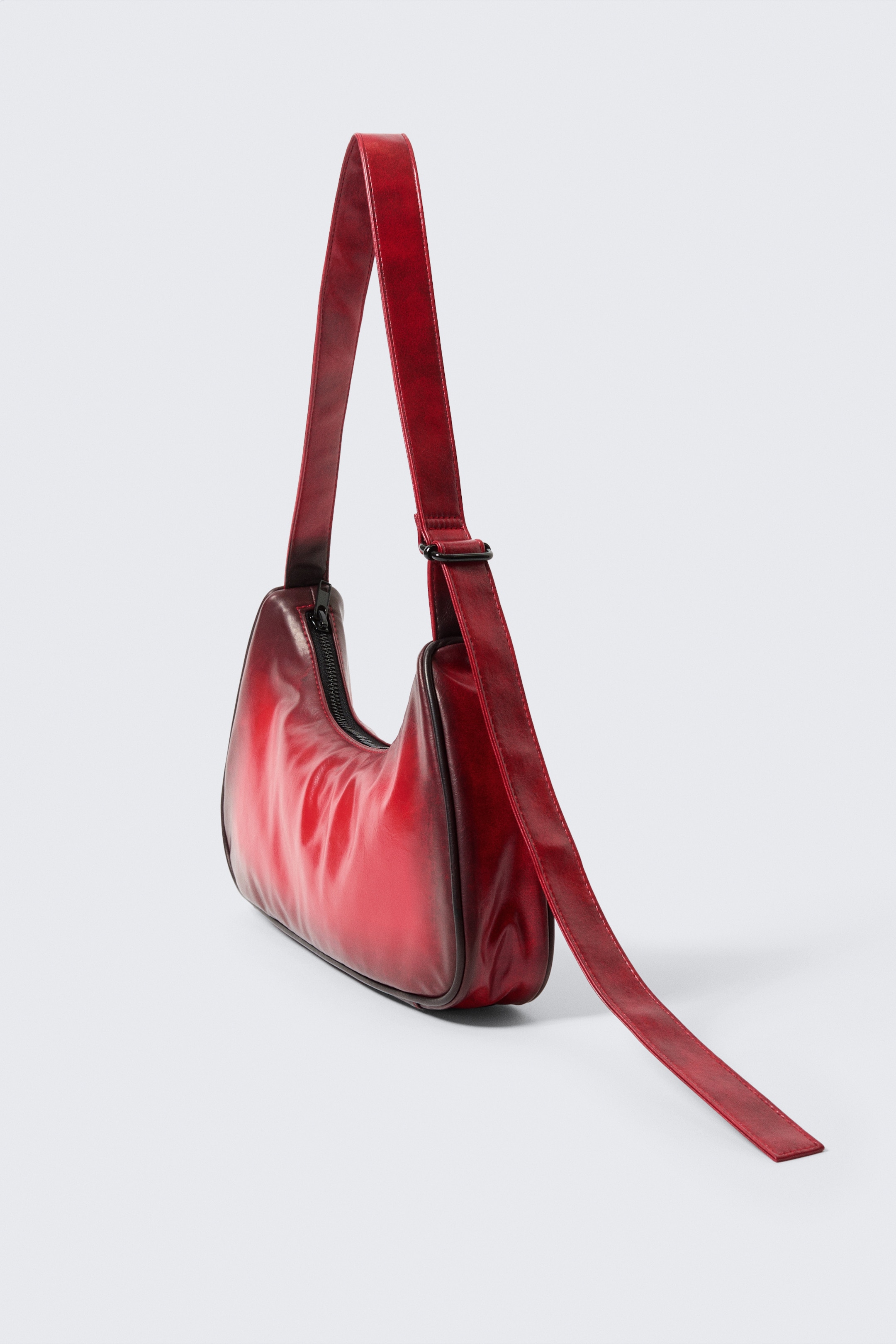 Agrandir l'image: Un sac à main rouge affiche une silhouette douce et sculptée avec une finition dégradée qui s'assombrit vers ses bords. Il est doté d'une fermeture zippée sur le dessus et d'une large sangle qui s'ajuste via une boucle en métal noir, se terminant par un détail de longue queue.