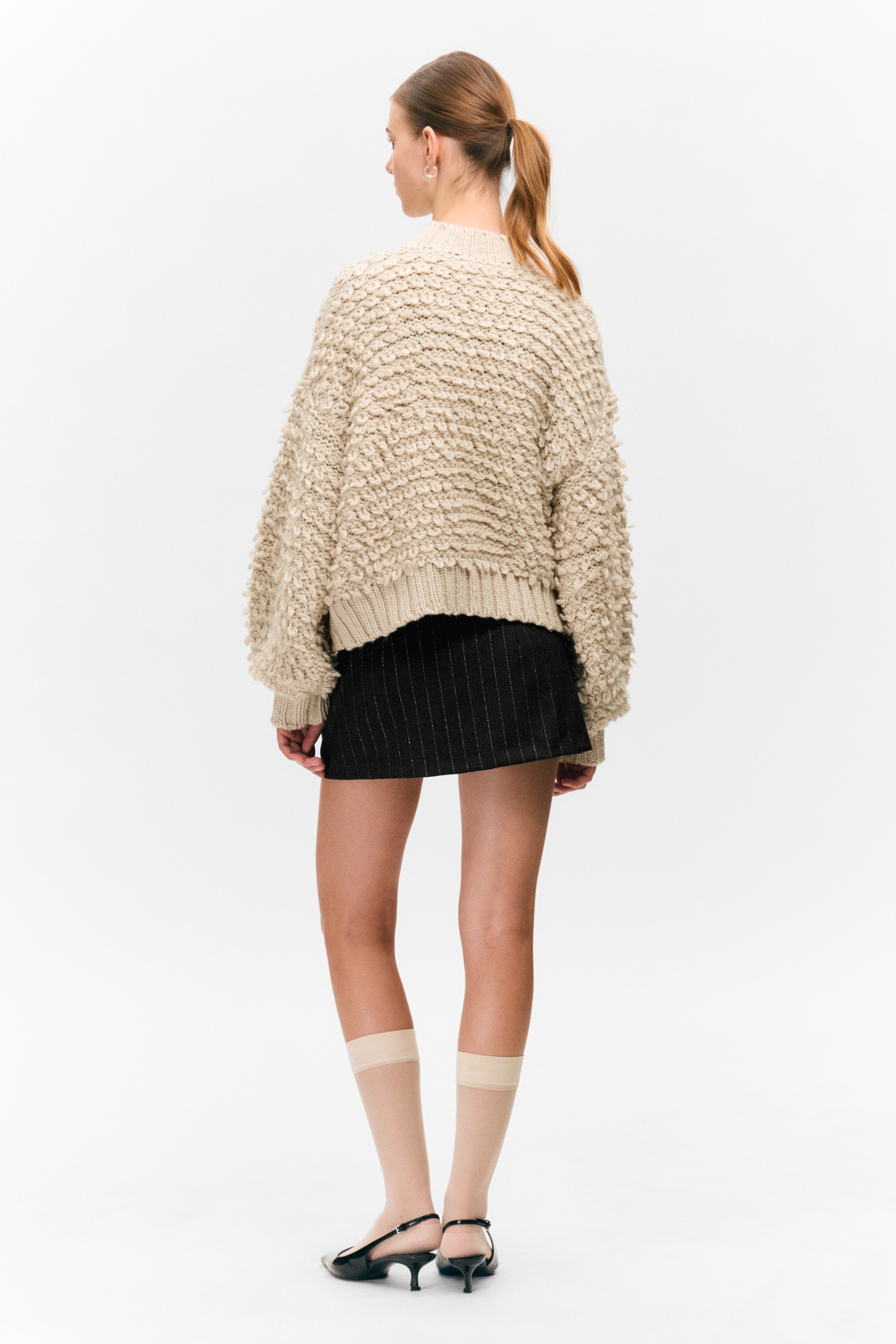 View larger image: A beige, textured knit sweater, appearing oversized and cropped, is being shown from the back, featuring a prominent bouclé-like texture and a ribbed hem. A black mini skirt with subtle vertical pinstripes is being worn beneath the sweater, visible below its hem.