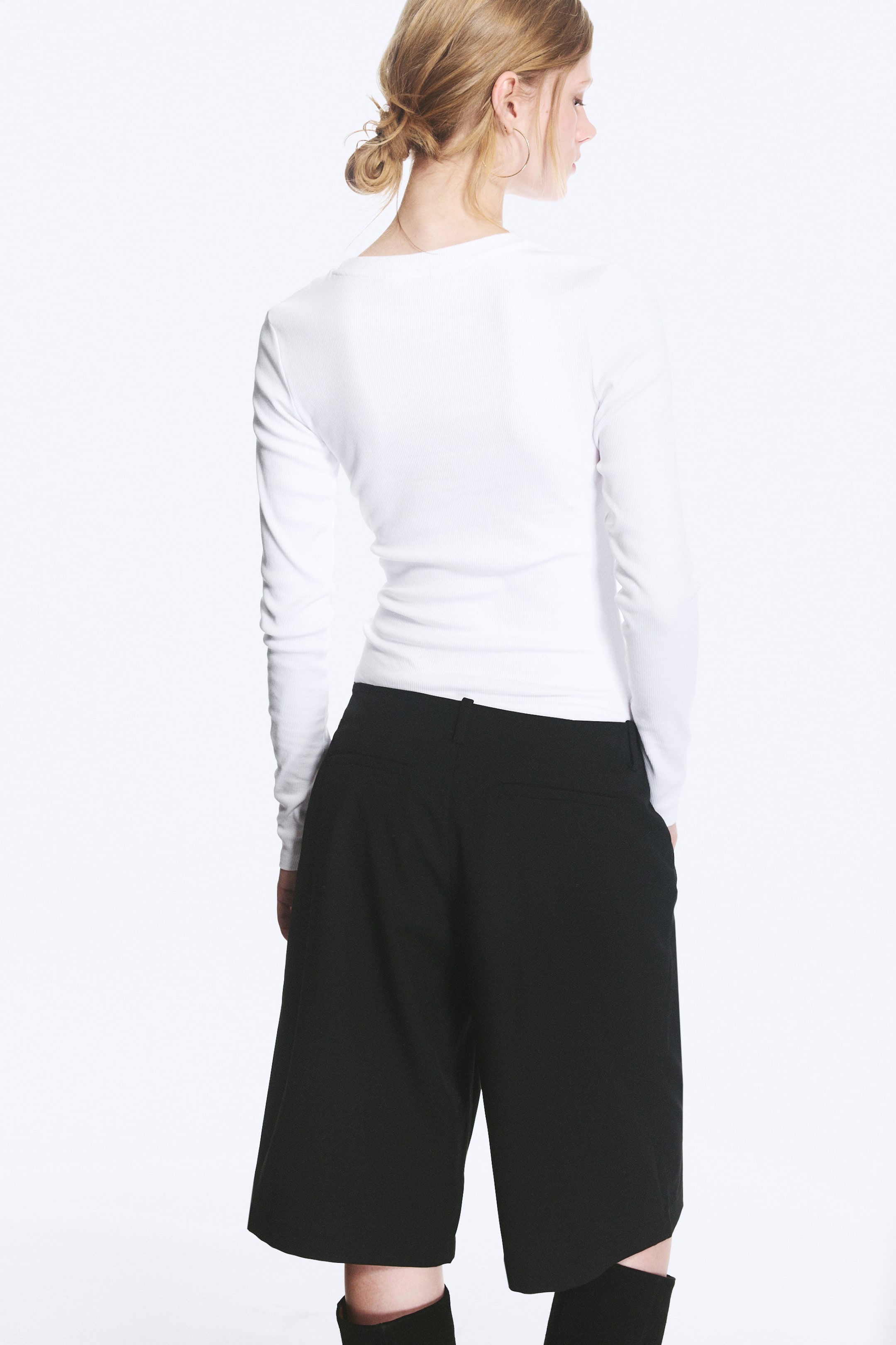Agrandir l'image: Le modèle porte un haut blanc à manches longues et à côtes serrées. Il présente un décolleté simple dans le dos et est rentré dans un pantalon culotte noir.