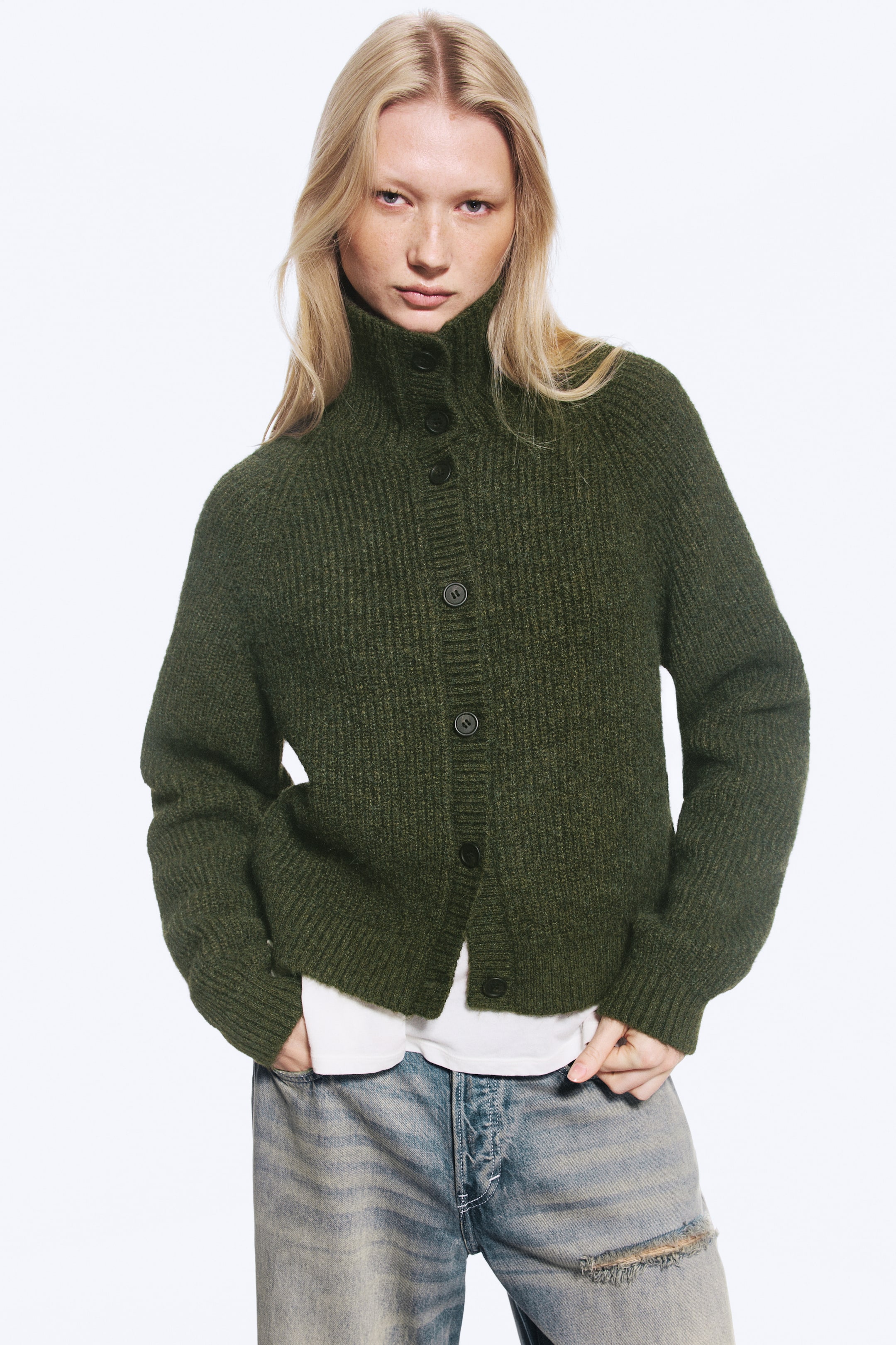 Agrandir l'image: Un cardigan en maille côtelée vert foncé avec un col montant boutonné est porté par le mannequin. Le cardigan est superposé à un t-shirt blanc et associé à un jean bleu délavé.